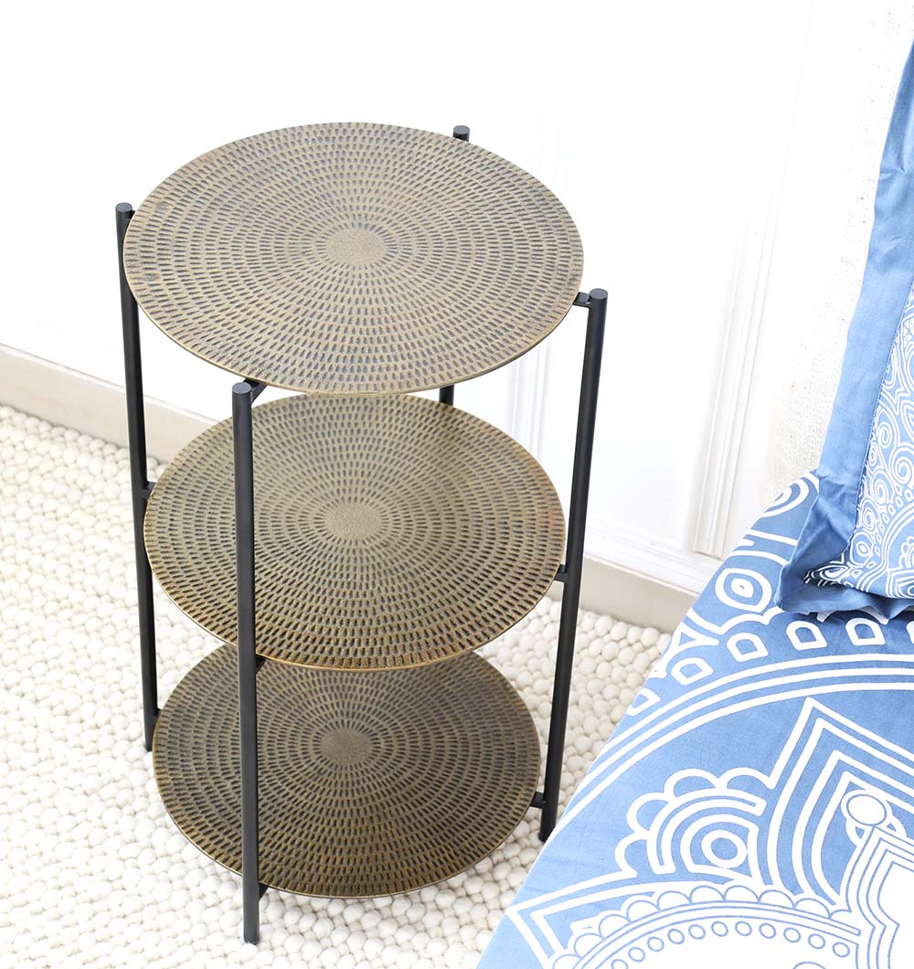 The RAMSHA HOME 3-Tier Round Side Tables (GH-075) feature an antique brass finish with patterned gold shelves and a black iron base, placed beside a blue and white patterned couch on a pebble-textured floor against a white wall.