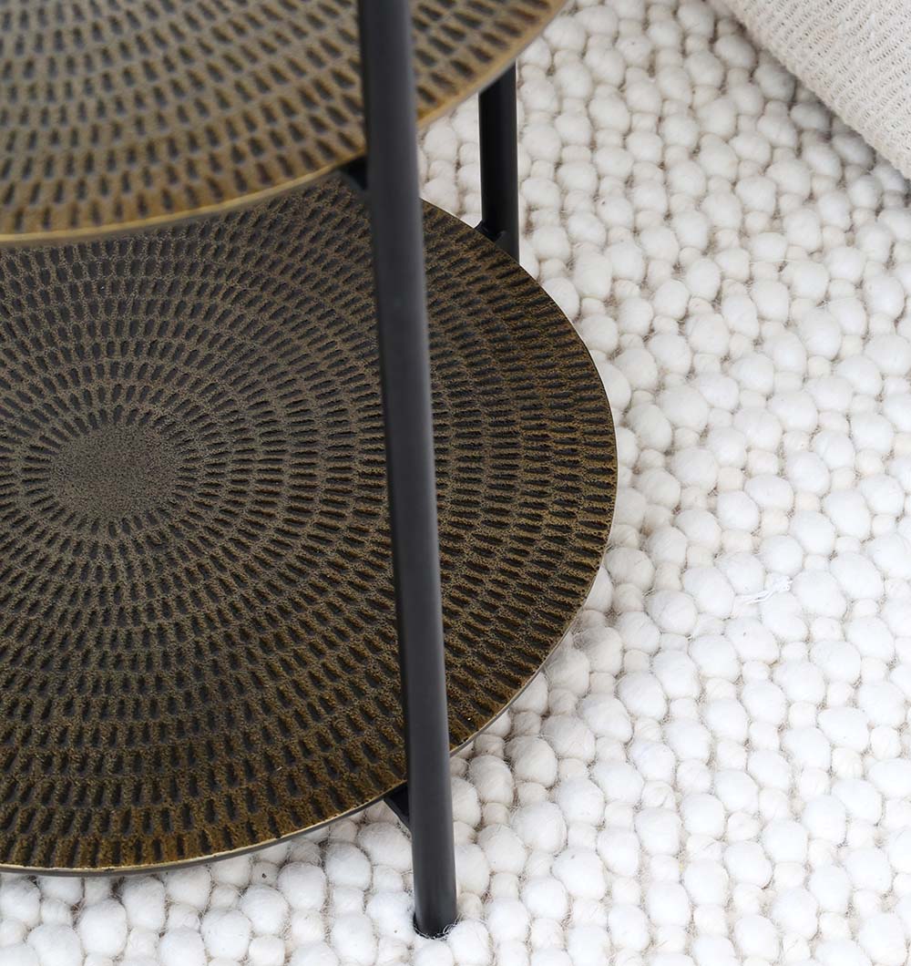 Close-up of RAMSHA HOME’s 3-Tier Round Side Tables (GH-075) featuring an antique brass finish and black iron base, placed on a thick white woven rug. Its elegant, patterned surface adds timeless charm to any space.