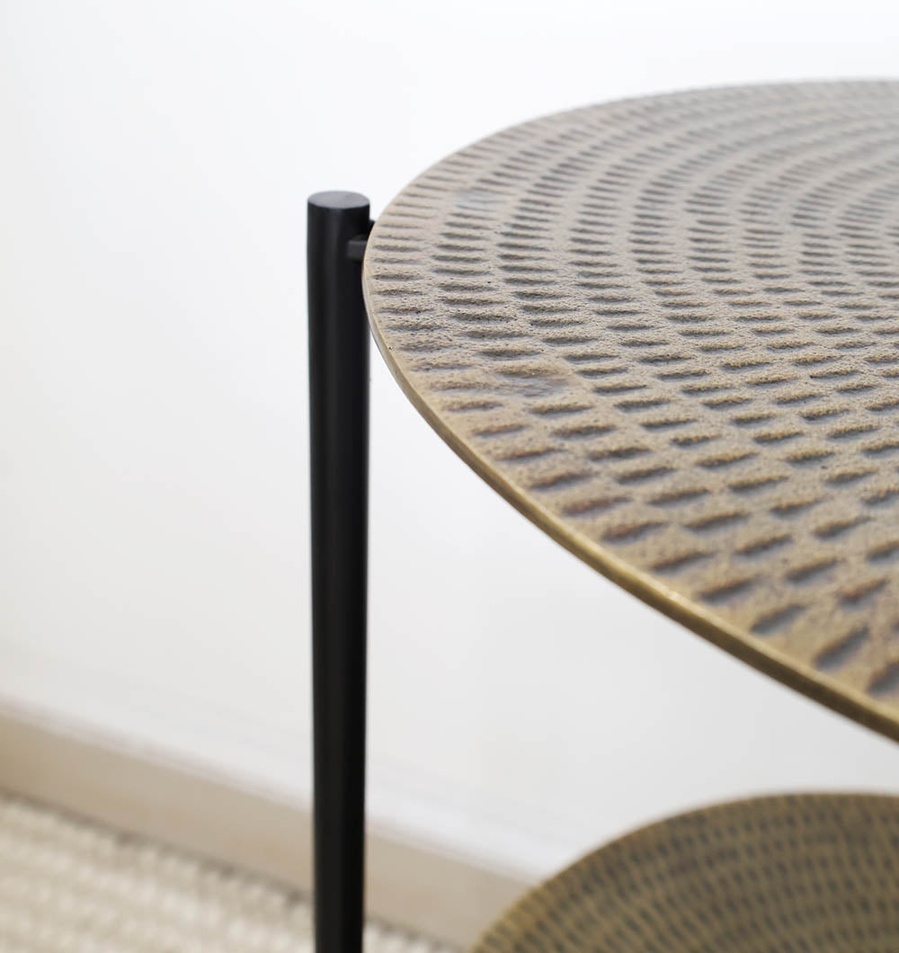 Close-up of RAMSHA HOME’s GH-075 3-Tier Round Side Table featuring a textured, patterned surface and antique brass finish. The black iron base supports tiered shelves, with a lower shelf partially visible against a light background.