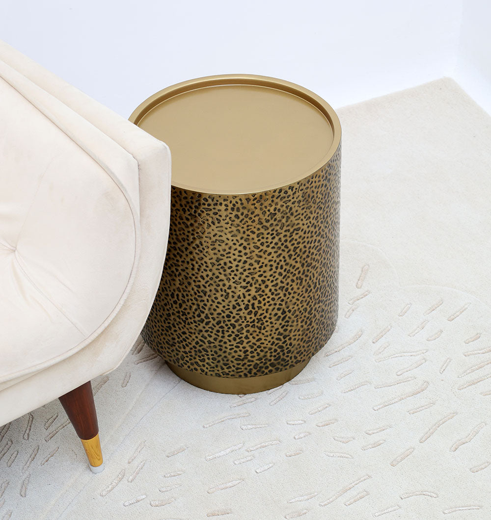 The RAMSHA HOME Luxury Iron Side Table – Golden Finish with Textured Design (GH-085) stands beside a beige upholstered chair with wooden legs on a light textured rug.