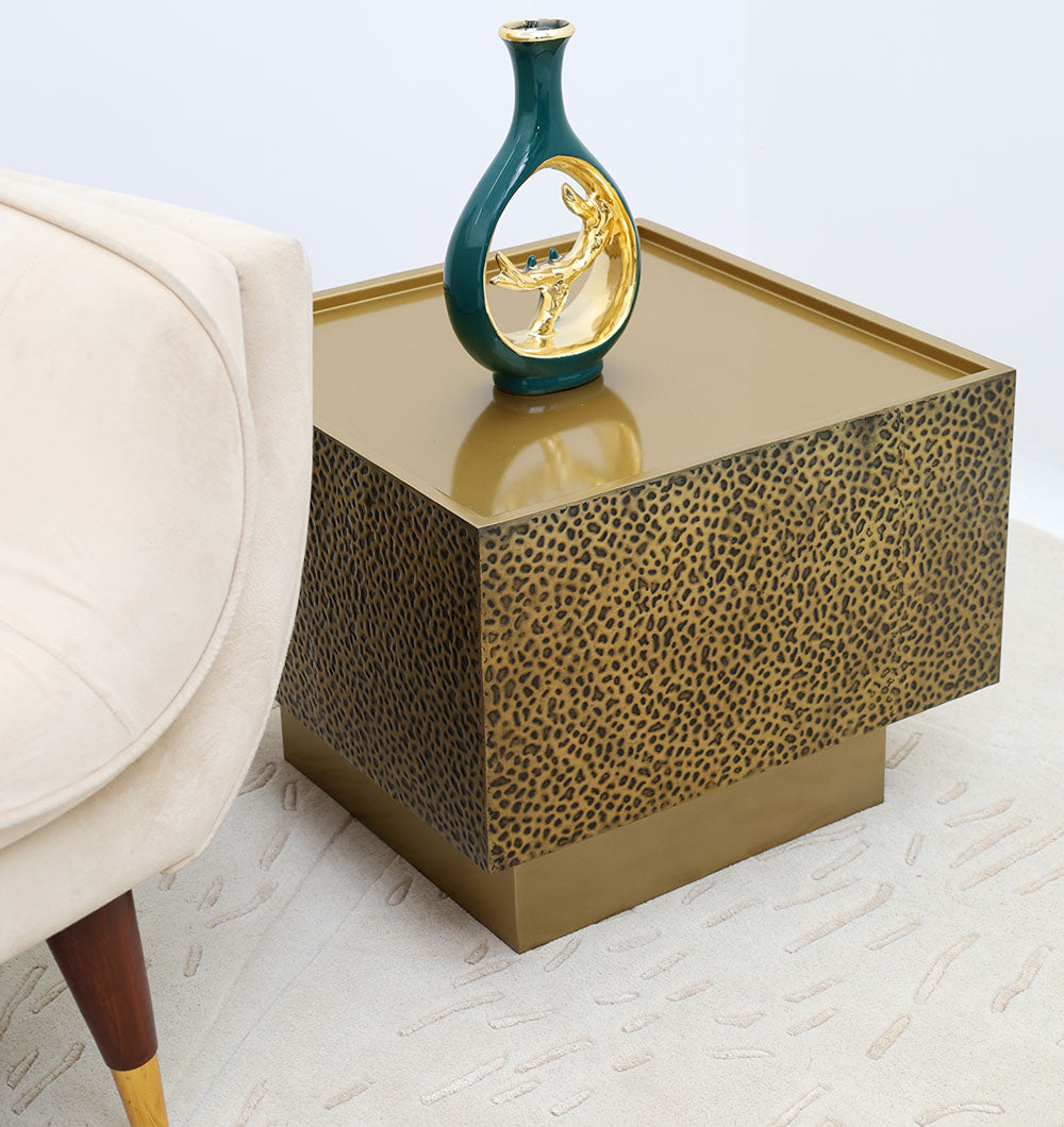 The RAMSHA HOME Luxury Square Iron Side Table – Golden Finish with Textured Design (GH-086) features a glossy brass antique look. It holds a small green and gold vase and sits beside a cream upholstered chair on a patterned light rug.
