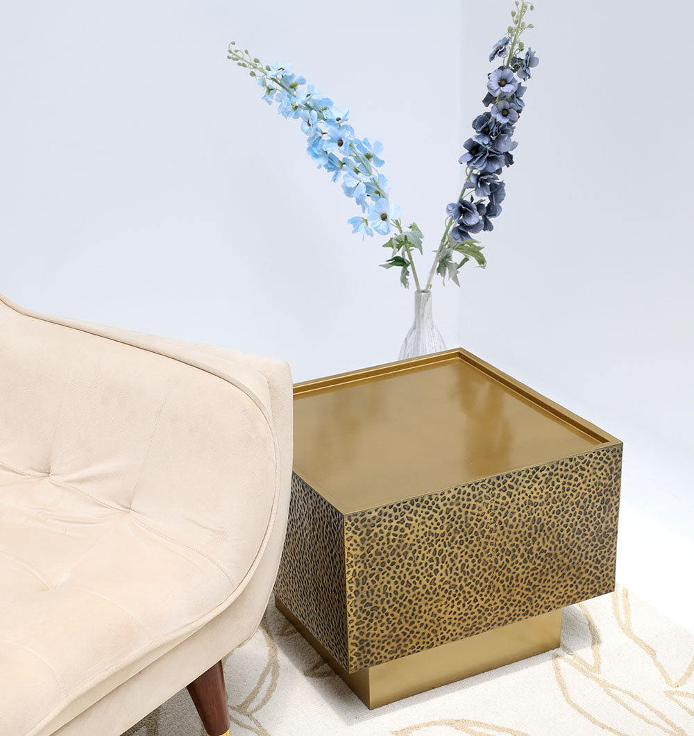 The RAMSHA HOME Luxury Square Iron Side Table (GH-086) with a golden finish and textured design sits next to a beige upholstered chair. Two tall artificial blue flowers in a clear vase add style—ideal for modern spaces.