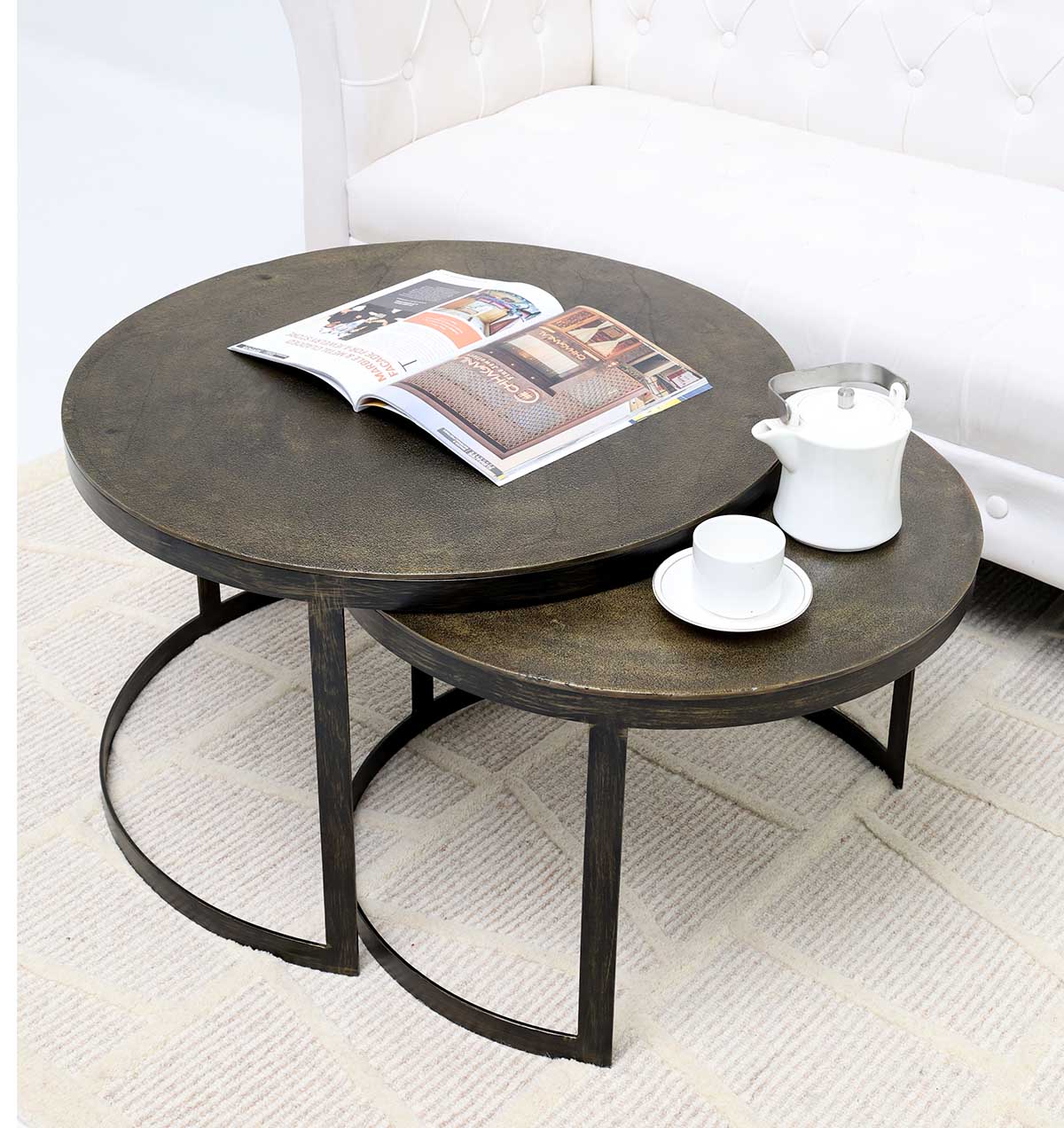 The RAMSHA HOME Modern Nesting Coffee Table Set (GH-089) features two round wooden-top tables with dark metal frames on a patterned rug. The larger holds an open magazine; the smaller displays a teapot, cup, and saucer. A white tufted sofa sits behind.