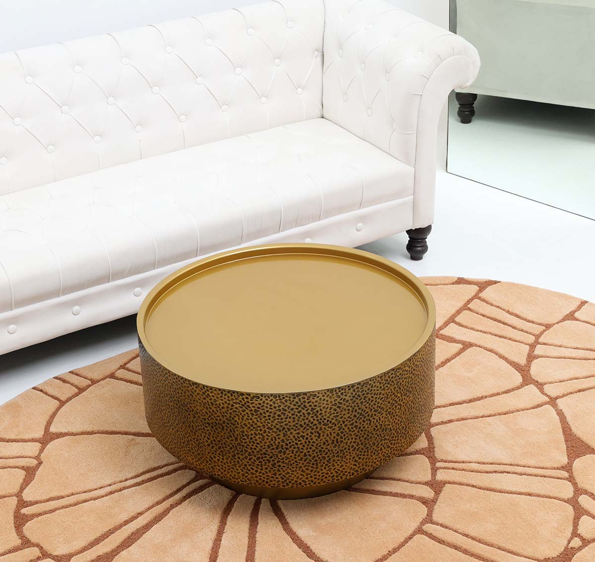 The RAMSHA HOME Luxury Round Iron Side Table – Golden Finish with Textured Design GH-090 stands on a patterned rug in front of a white tufted sofa, enhancing the brightness of this modern living room.