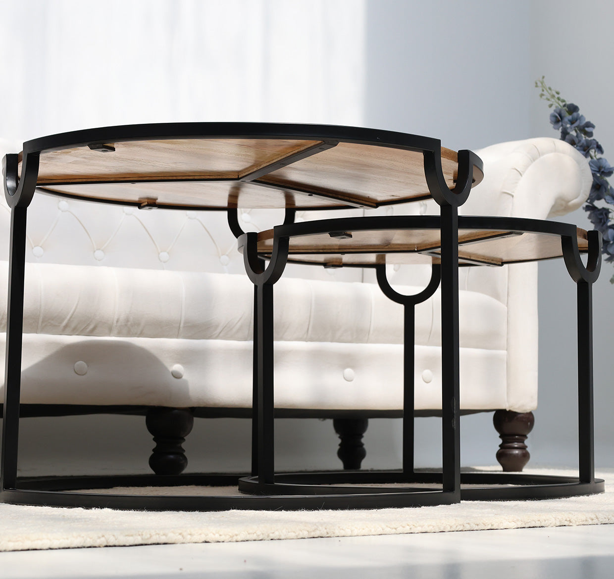 The RAMSHA HOME Modern Nesting Coffee Table Set (GH-094), with round wooden tops and black metal frames, sits in front of a white tufted sofa on a light rug. A small bunch of purple flowers rests on the sofa.