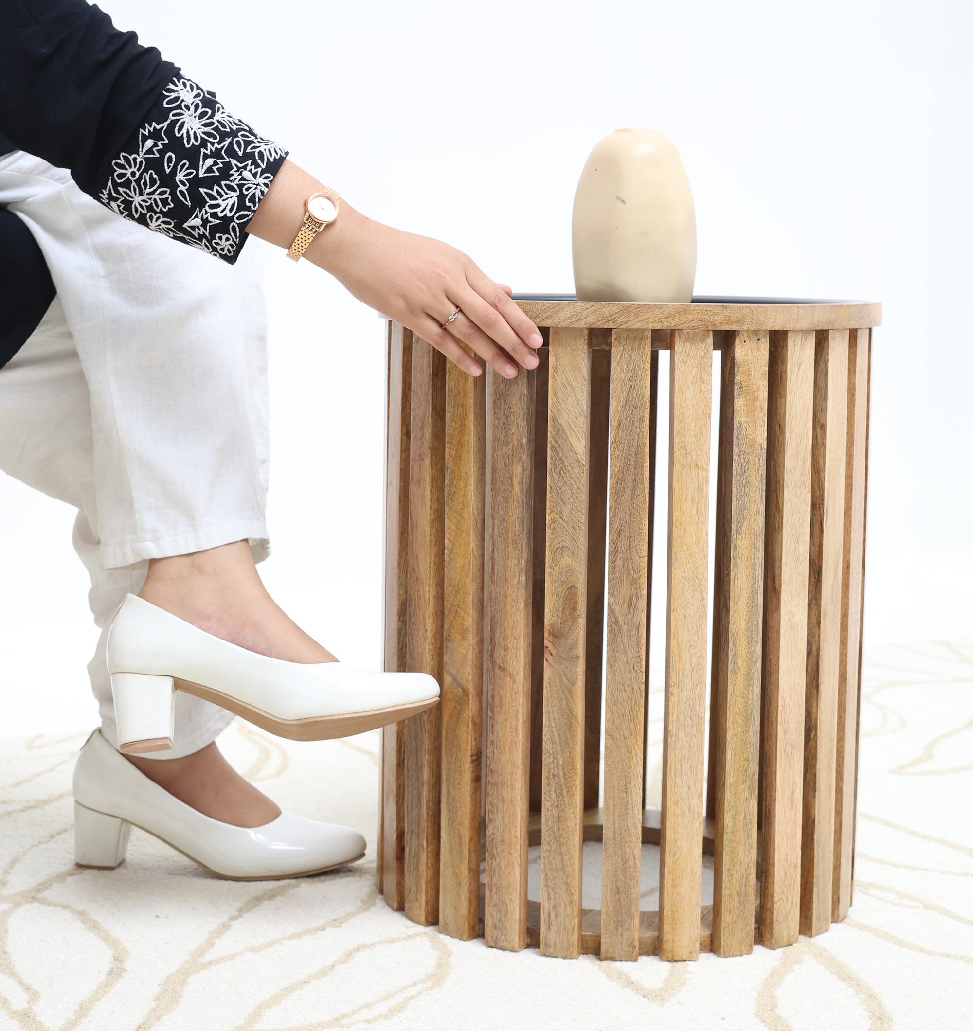 A person in white pants and heels sits beside the RAMSHA HOME Modern Wooden & Iron Coffee Table – Cylindrical Slatted Design GH-097, resting their hand near a small beige vase on top.