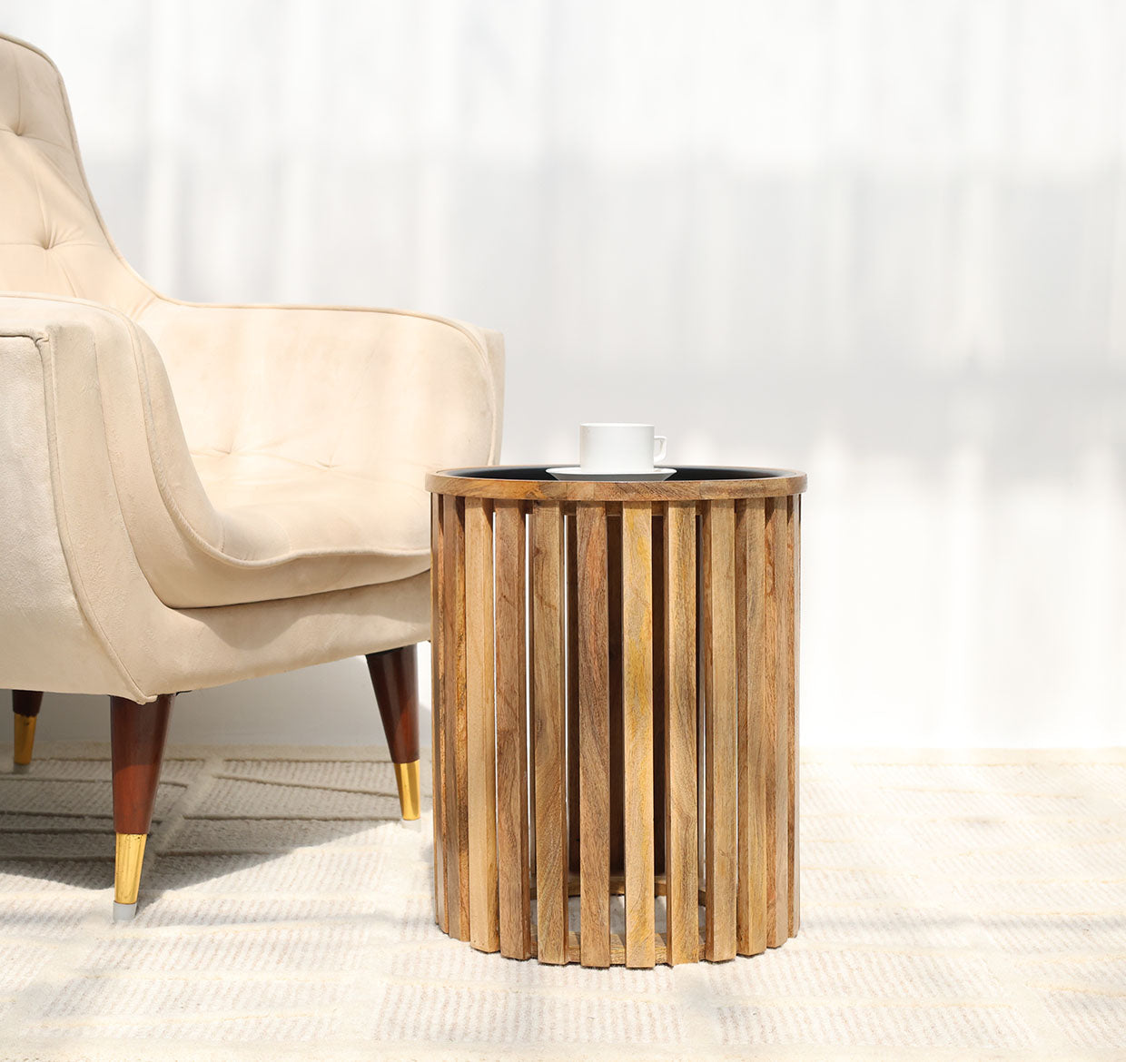 A beige armchair with wooden legs sits beside the RAMSHA HOME Modern Wooden & Iron Coffee Table (Cylindrical Slatted Design GH-097), where a white cup and saucer rest, as sunlight casts shadows on the light floor and wall.