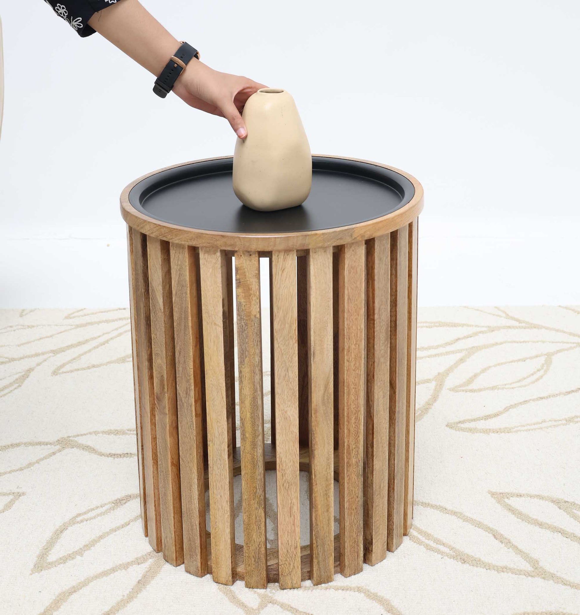 A person places a beige vase on the RAMSHA HOME Modern Wooden & Iron Coffee Table – Cylindrical Slatted Design GH-097, which has vertical wooden slats and a black top, set on a light leaf-patterned carpet.