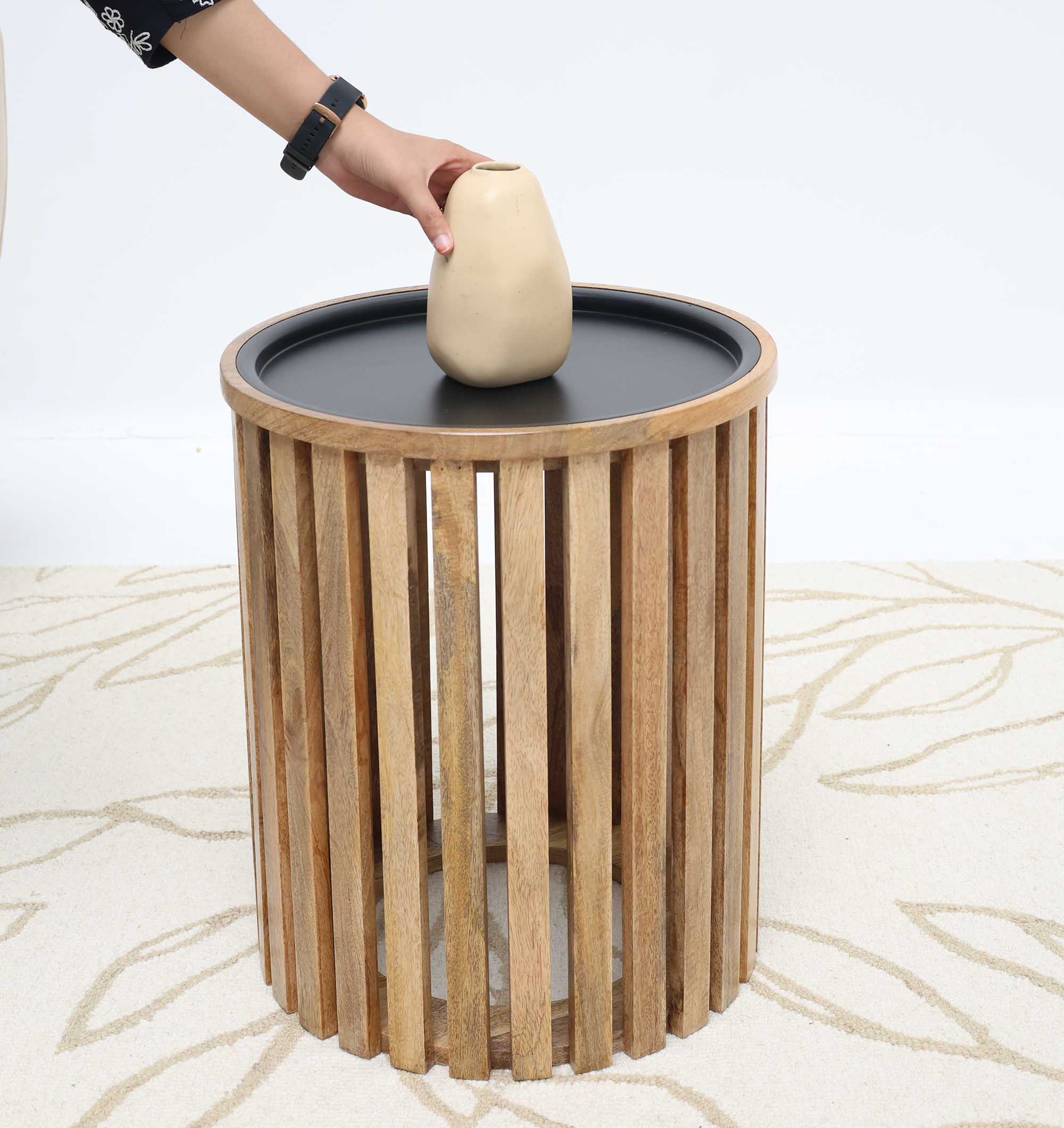 A person places a beige vase on the RAMSHA HOME Modern Wooden & Iron Coffee Table – Cylindrical Slatted Design GH-097, which has vertical wooden slats and a black top, set on a light leaf-patterned carpet.
