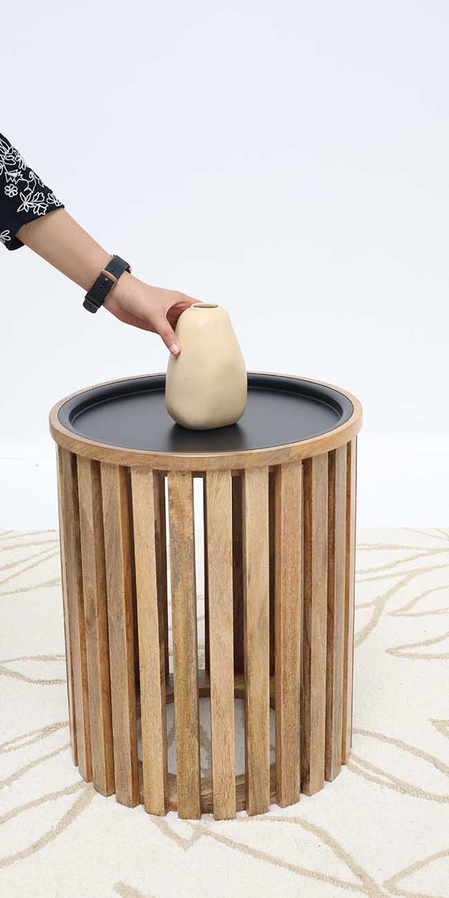 A hand places a beige vase on the RAMSHA HOME Modern Wooden & Iron Coffee Table – Cylindrical Slatted Design GH-097, set atop a light-patterned rug with a plain white background.