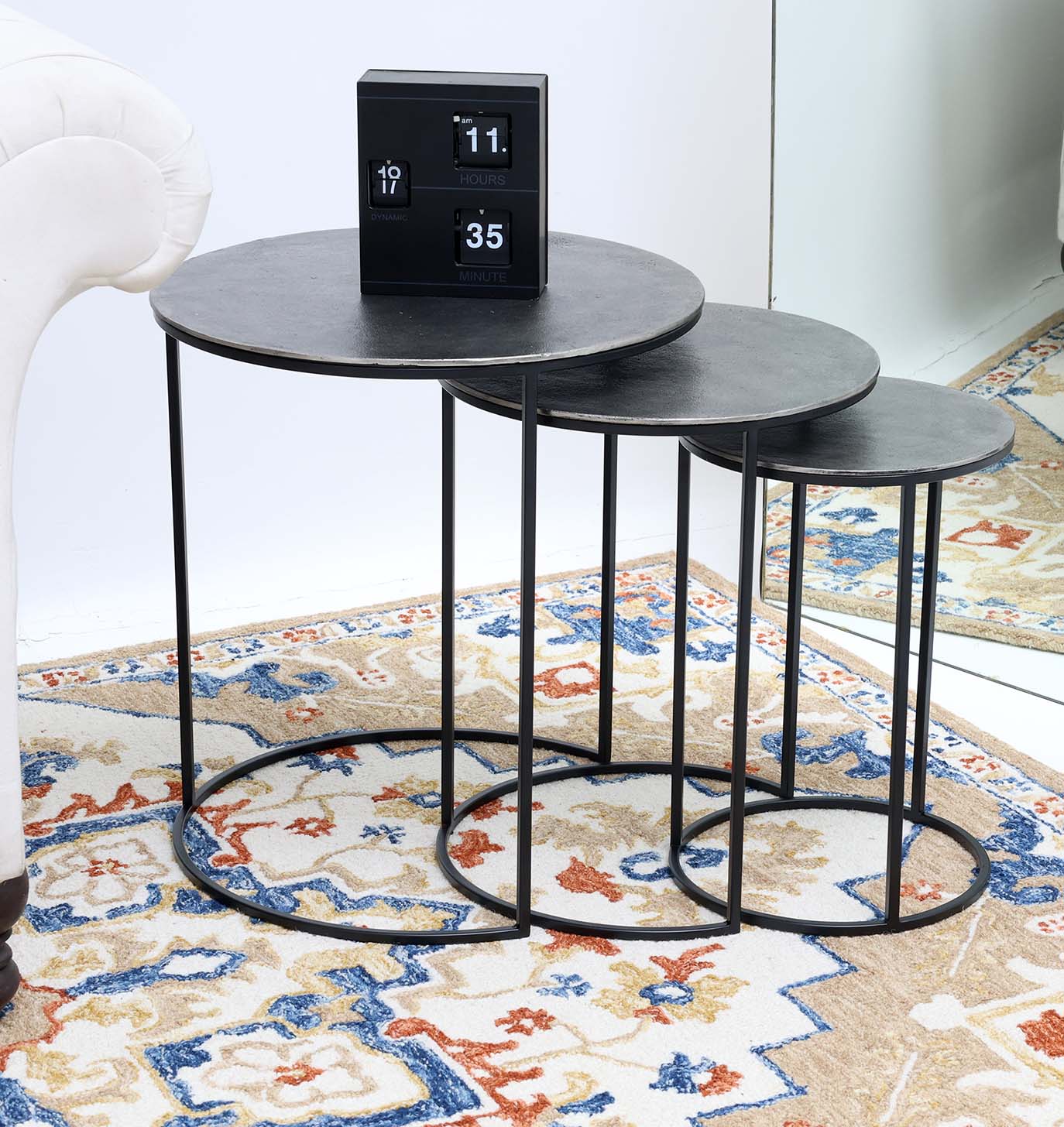 The RAMSHA HOME Set of 3 Modern Nesting Side Tables (Antique Brass Finish, Black Iron Base GH-071) are arranged by a white sofa on a patterned rug. A black flip clock adds modern style, with a mirror in the background.