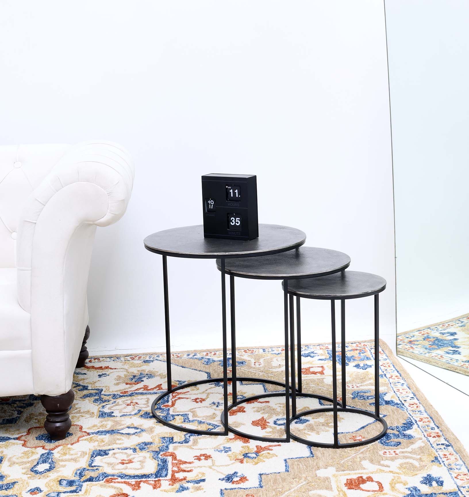 A RAMSHA HOME Set of 3 Modern Nesting Side Tables (Antique Brass Finish, Black Iron Base, GH-071) sits by a white sofa and patterned rug. A black flip clock is on the largest table, with a mirror on the right.