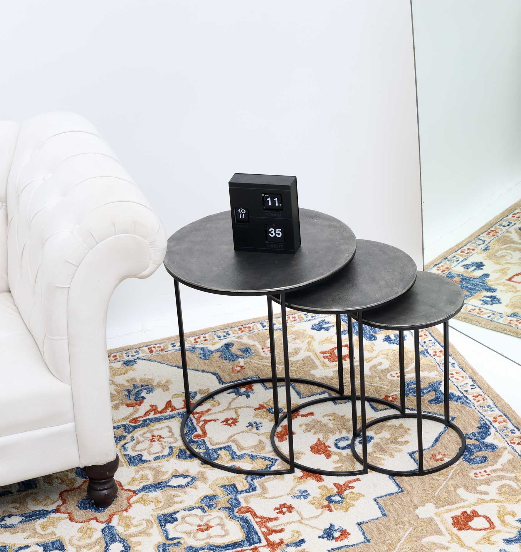 The RAMSHA HOME Set of 3 Modern Nesting Side Tables (Antique Brass Finish, Black Iron Base, GH-071) sits beside a white sofa on a patterned rug, topped with a digital clock, while a large mirror reflects the stylish arrangement.