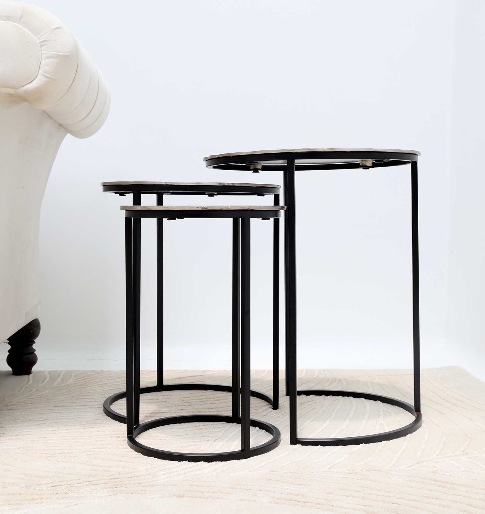 A set of 3 modern nesting side tables by RAMSHA HOME, featuring antique brass tops and grey iron bases (GH-111-A), sits on a light rug next to a white upholstered sofa in a minimal, bright room.