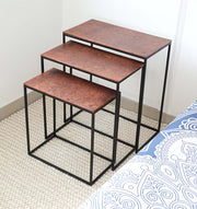 The RAMSHA HOME Modern Three-Tier Side Tables (GH-111-B) with black powder/copper finish and black iron base are stacked by size on a light textured rug beside a bed with blue and white patterned bedding.