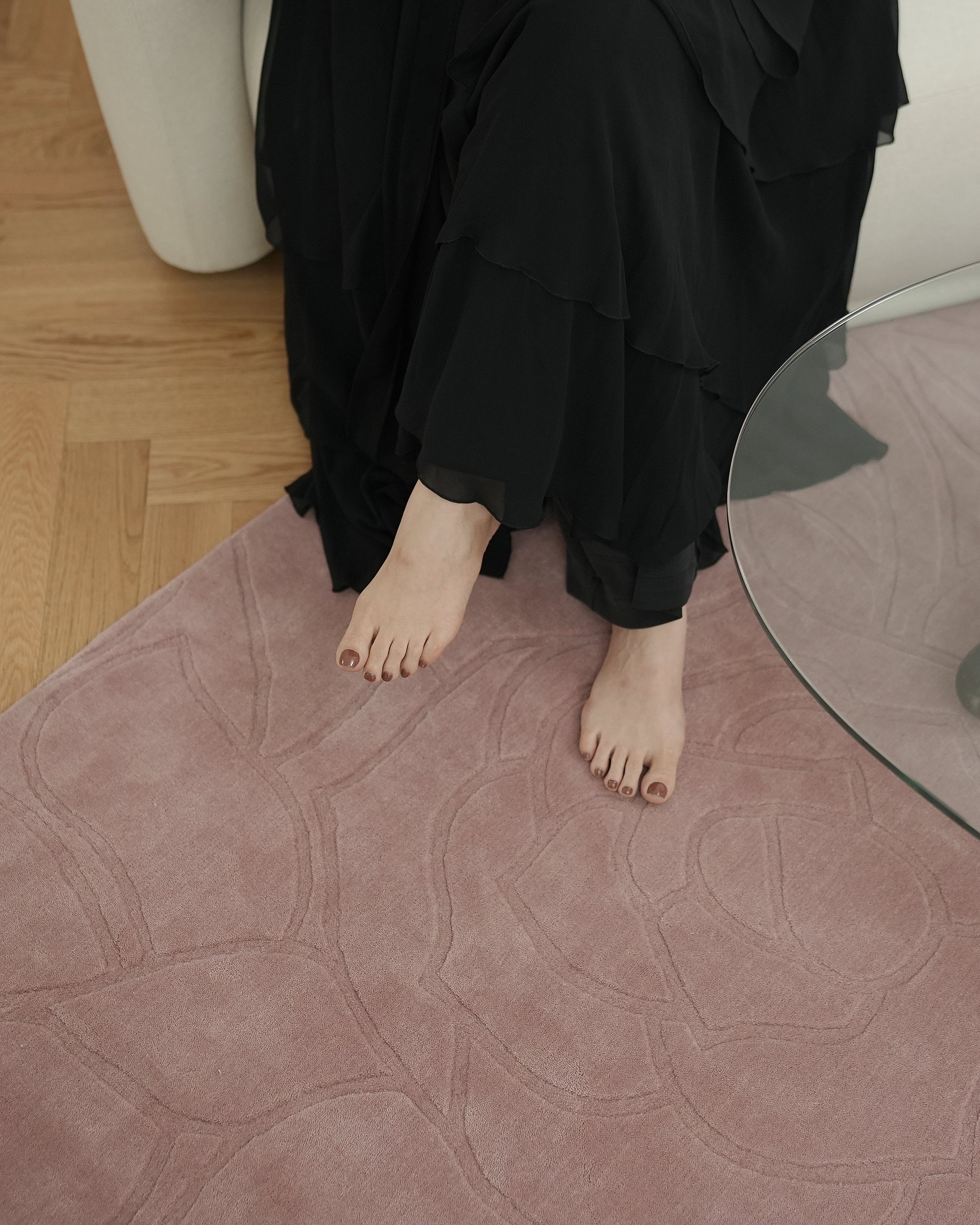 A person in a long black ruffled dress sits on a sofa with bare feet resting on the RAMSHA HOME Rectangle Handmade Pink Color Area Rug - LKH-029, placed beside a glass coffee table on a wooden floor.