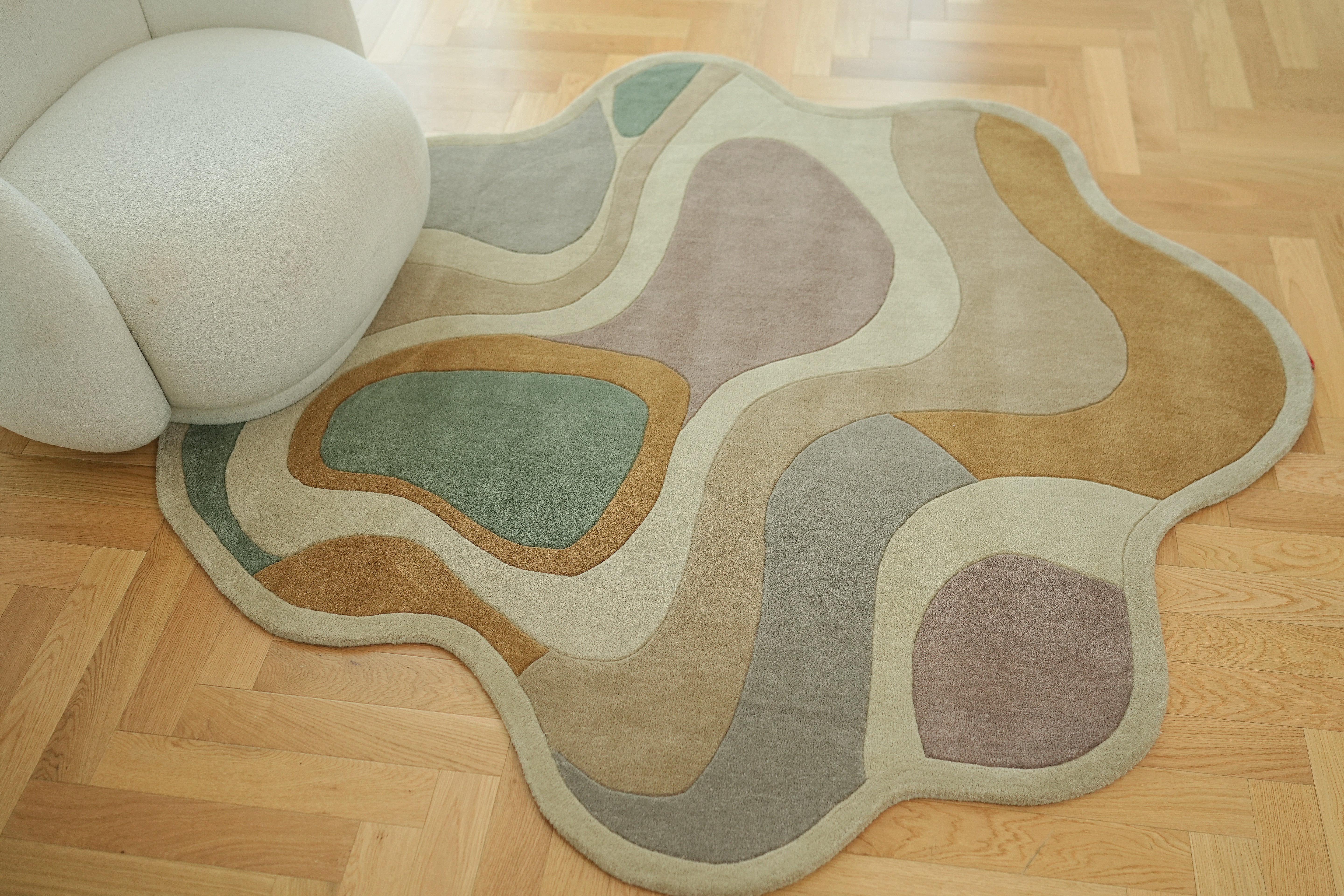 The RAMSHA HOME Handmade Multicolor Shaped Area Rug HTW 127 (200cm) in green, beige, brown, and gray rests on a wooden parquet floor beside a curved off-white chair.
