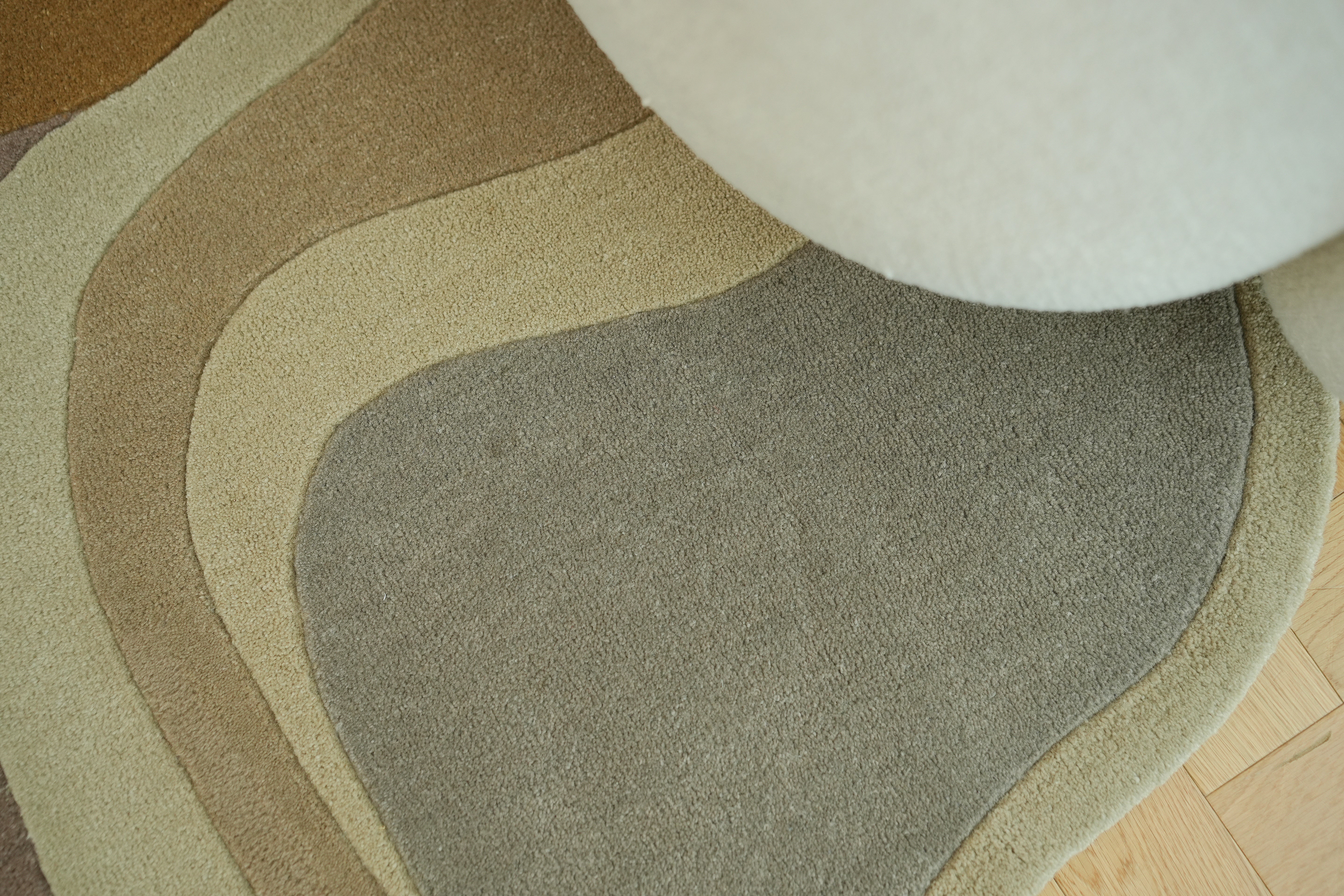 A close-up of the RAMSHA HOME Handmade Multicolor Shaped Area Rug HTW 127 (200cm), featuring beige, tan, and gray wavy patterns, partially covered by a rounded light upholstered piece on a wooden floor.