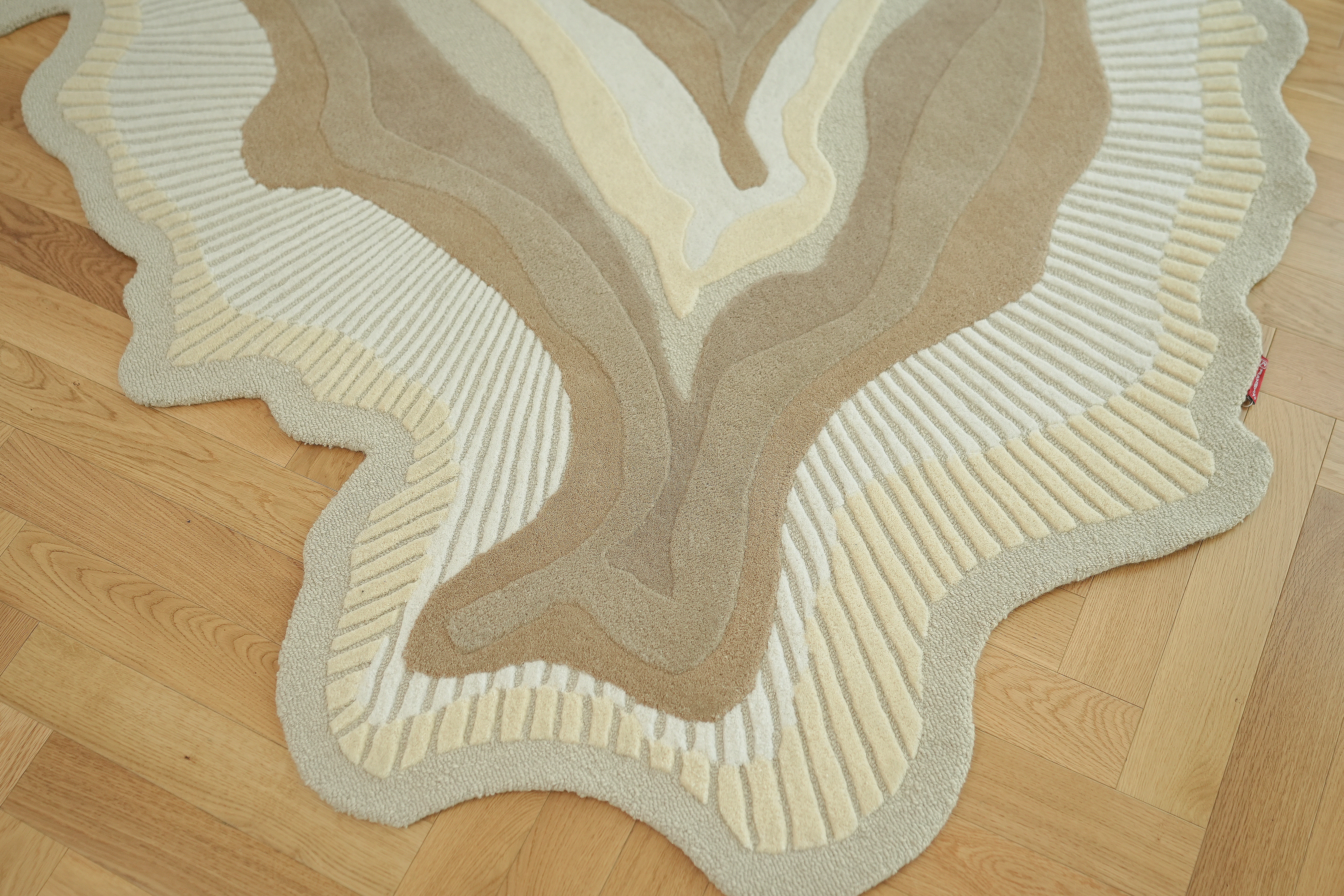 The RAMSHA HOME Handmade Shape Wool & Cotton Earth Tone Rug HTW 124 (200cm), featuring abstract wavy patterns in beige and cream, lies on a herringbone-patterned wooden floor.