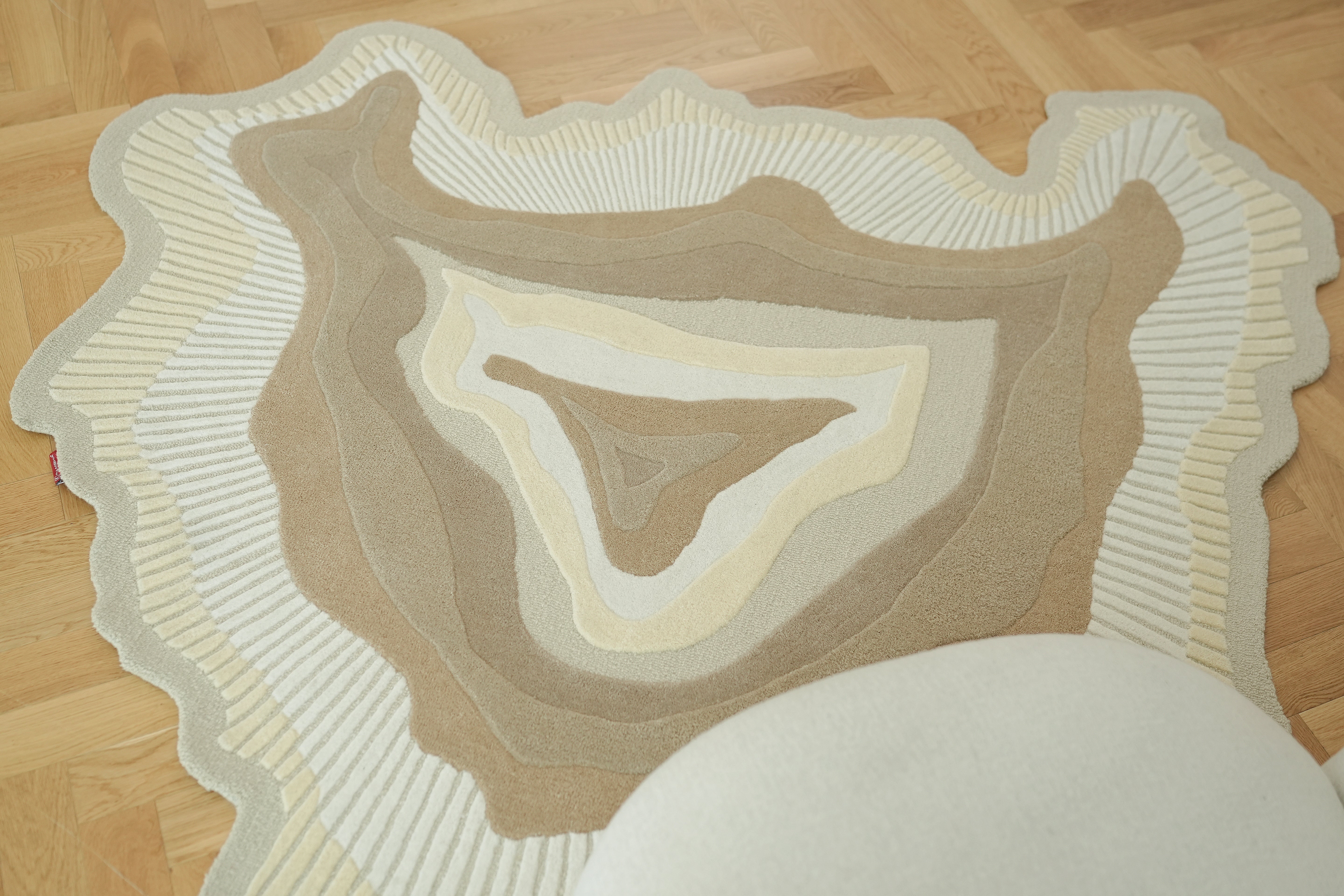 The RAMSHA HOME Handmade Shape Wool & Cotton Earth Tone Rug HTW 124 (200cm), with beige, cream, and brown wavy patterns, lies on a light wood parquet floor. A round, light-colored object is partially visible at its edge.