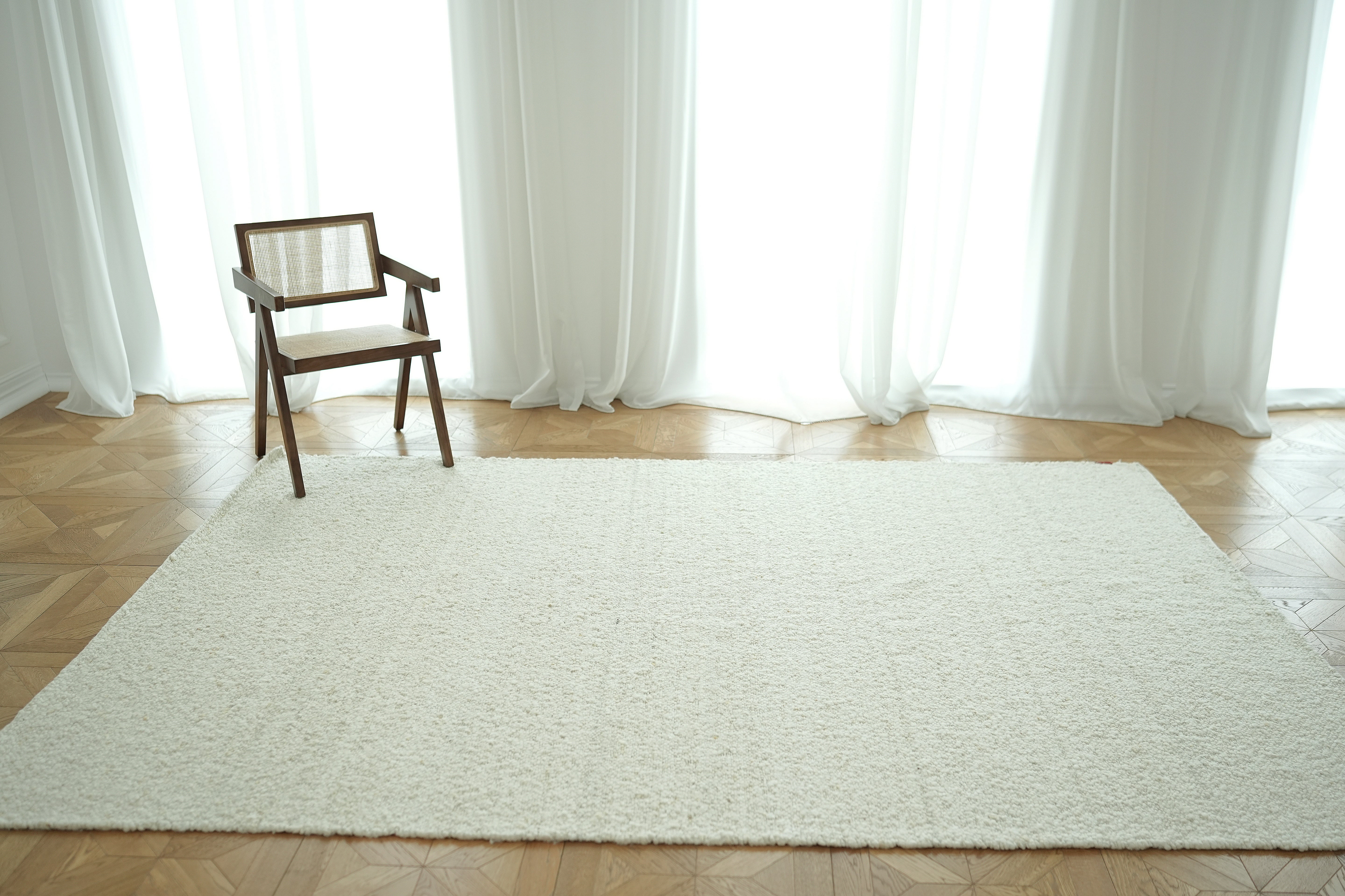 A single wooden chair with a cane backrest sits on the left side of the RAMSHA HOME Handmade Natural Off White Woven Rug For Home Decor SR-059 in a bright, minimalist room with sheer white curtains and a wooden floor.