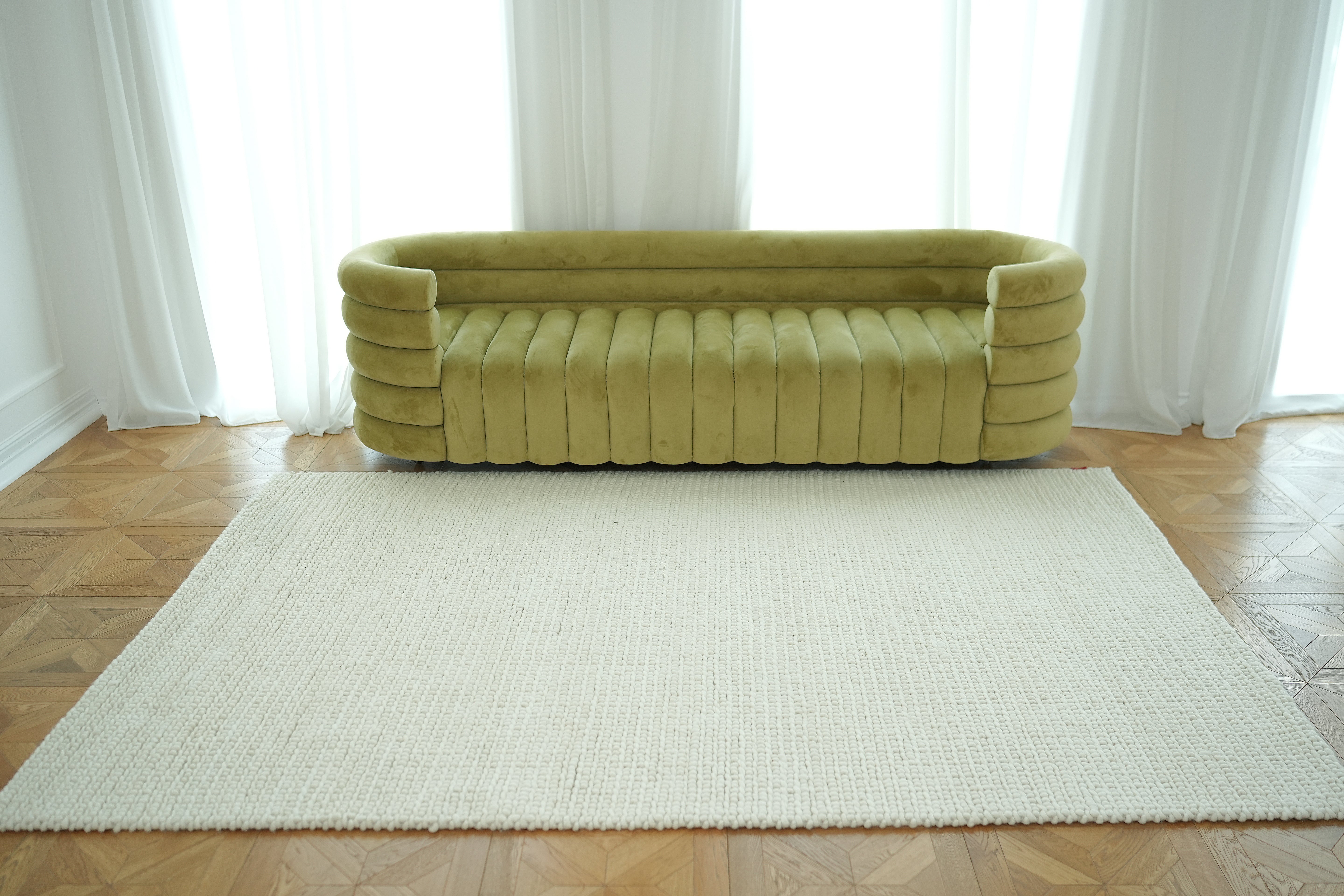 A modern olive green velvet sofa with rounded arms sits by white curtains and large windows, with a Handmade Natural White Woven Rug For Home Decor SRN-031 placed on the wooden parquet floor in front.