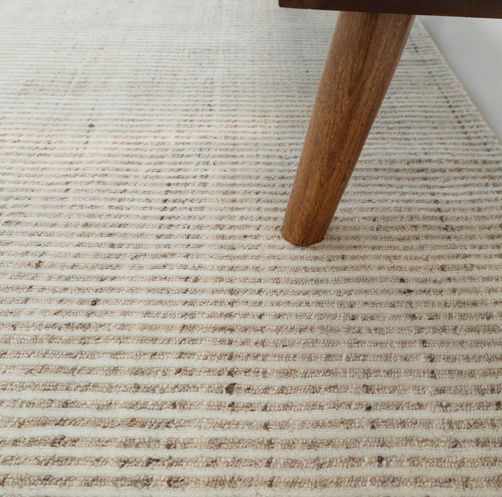 A close-up of RAMSHA HOME's Handwoven Ivory Wool Rug (HLR-001) in natural cream shows its textured, subtle horizontal stripes with a wooden furniture leg resting on this minimalist area rug.