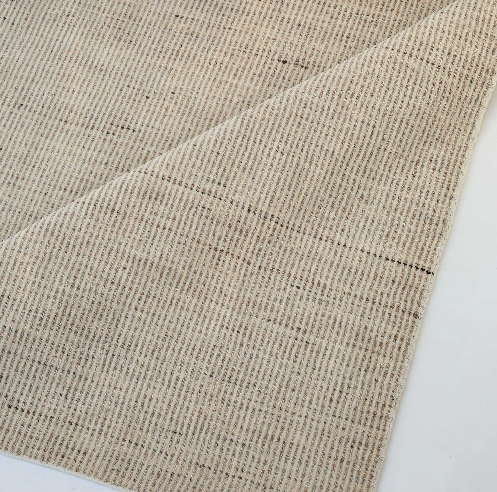 A close-up of the RAMSHA HOME Handwoven Ivory Wool Rug (HLR-001) in natural cream shows thin, evenly spaced brown stripes. One corner is partially folded to reveal the underside, with the minimalist rug against a white background.