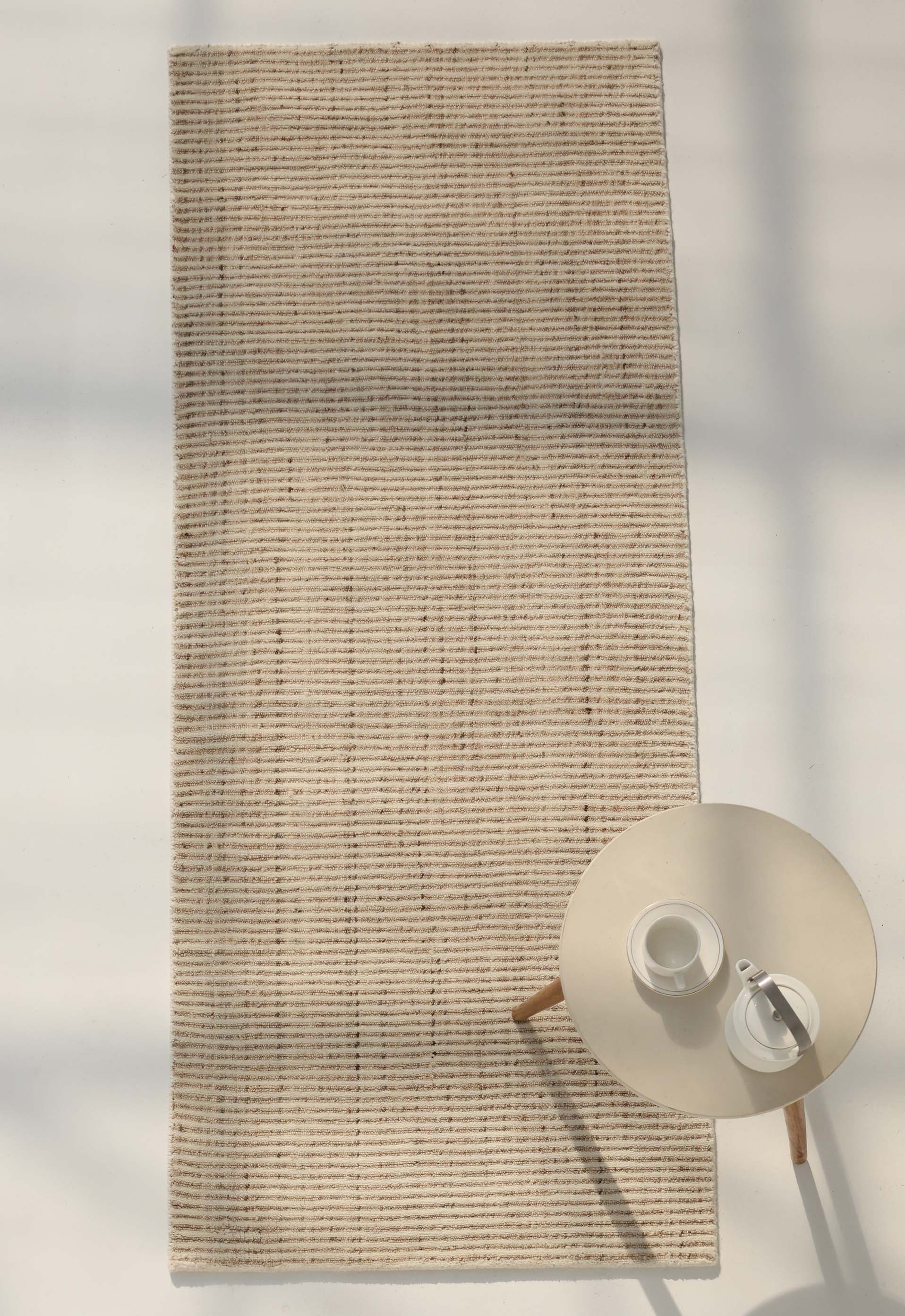 The RAMSHA HOME Handwoven Ivory Wool Rug (HLR-001), a rectangular area rug in natural cream, lies on a white floor. In the lower right corner, a small round table holds two white cups and saucers.
