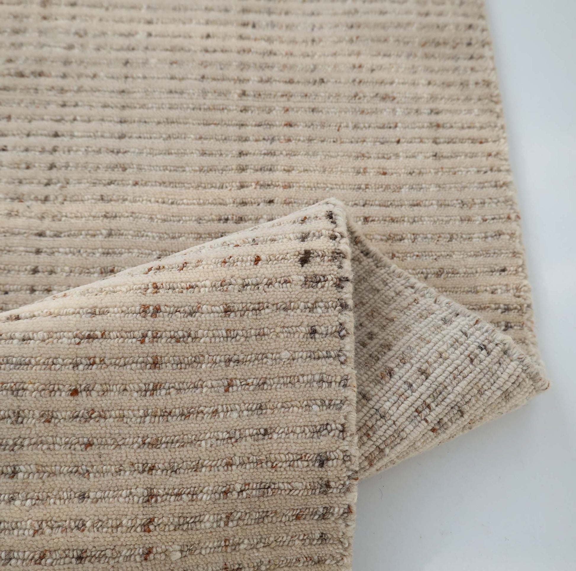 A close-up of the RAMSHA HOME Handmade Natural Wool Rug – Textured Beige Area Rug (HLR-002), featuring thin brown stripes, a folded corner, and a subtle speckled handwoven texture.
