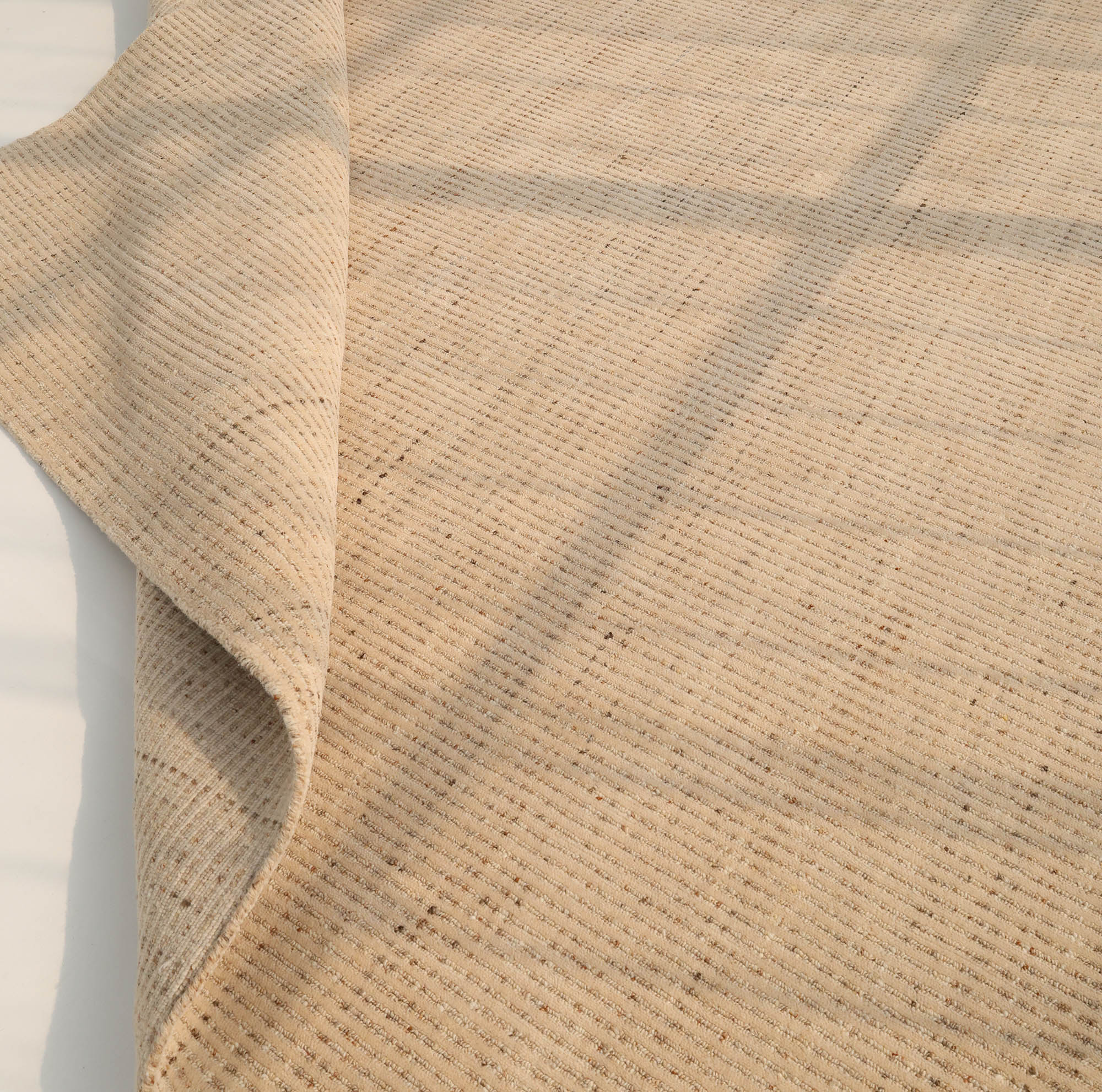A close-up of the RAMSHA HOME Handmade Natural Wool Rug – Textured Beige Area Rug (HLR-002) shows its subtle checkered texture and soft ivory hue, partially folded at the corner, resting on a light floor with gentle shadows.