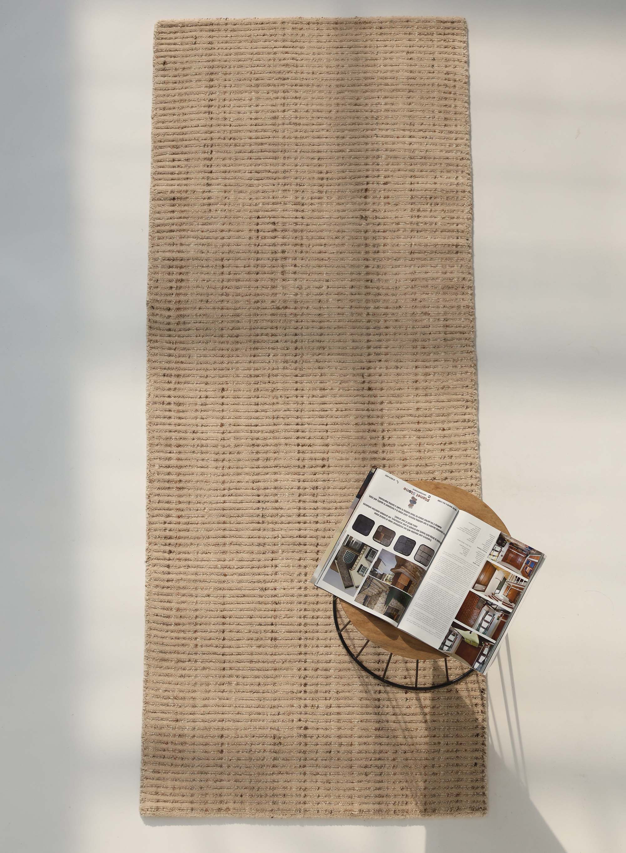 The RAMSHA HOME Handmade Natural Wool Rug – Textured Beige Area Rug (HLR-002) lies on a light floor. A round side table with an open decor magazine rests in one corner, where sunlight creates gentle shadows across the minimalist scene.