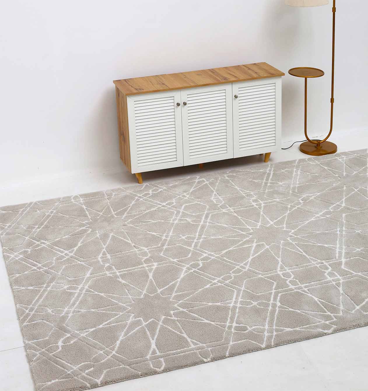 The RAMSHA HOME Decorative Rectangle Shape Rug HTP-15, light gray with a white geometric pattern, is placed on a white floor in front of a wooden cabinet with white slatted doors, beside a standing lamp with a round base.