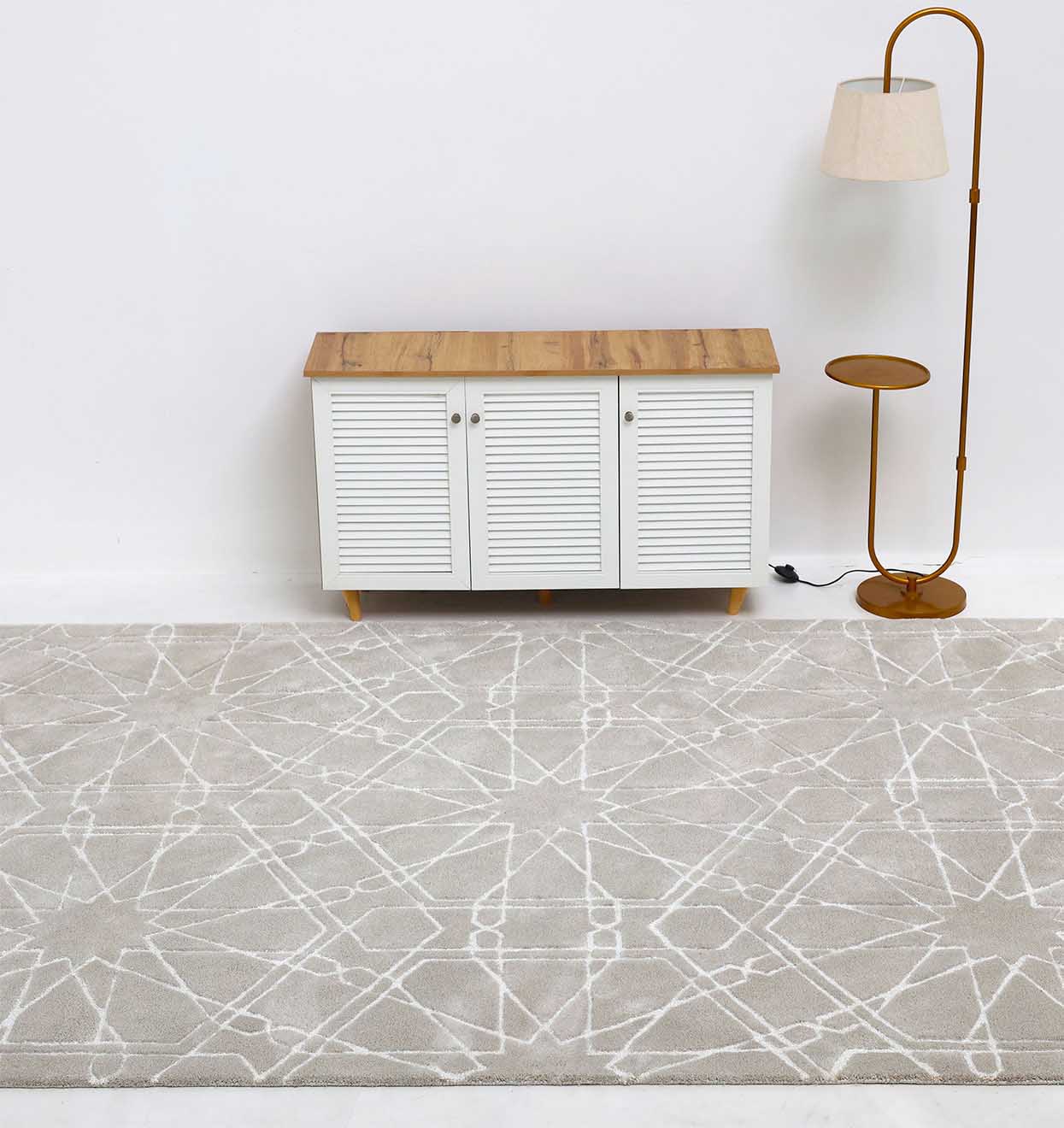 A modern living room featuring the RAMSHA HOME Decorative Rectangle Shape Rug HTP-15 in light gray with a white geometric pattern, paired with a white cabinet with wooden top and a tall gold floor lamp beside a round side table.