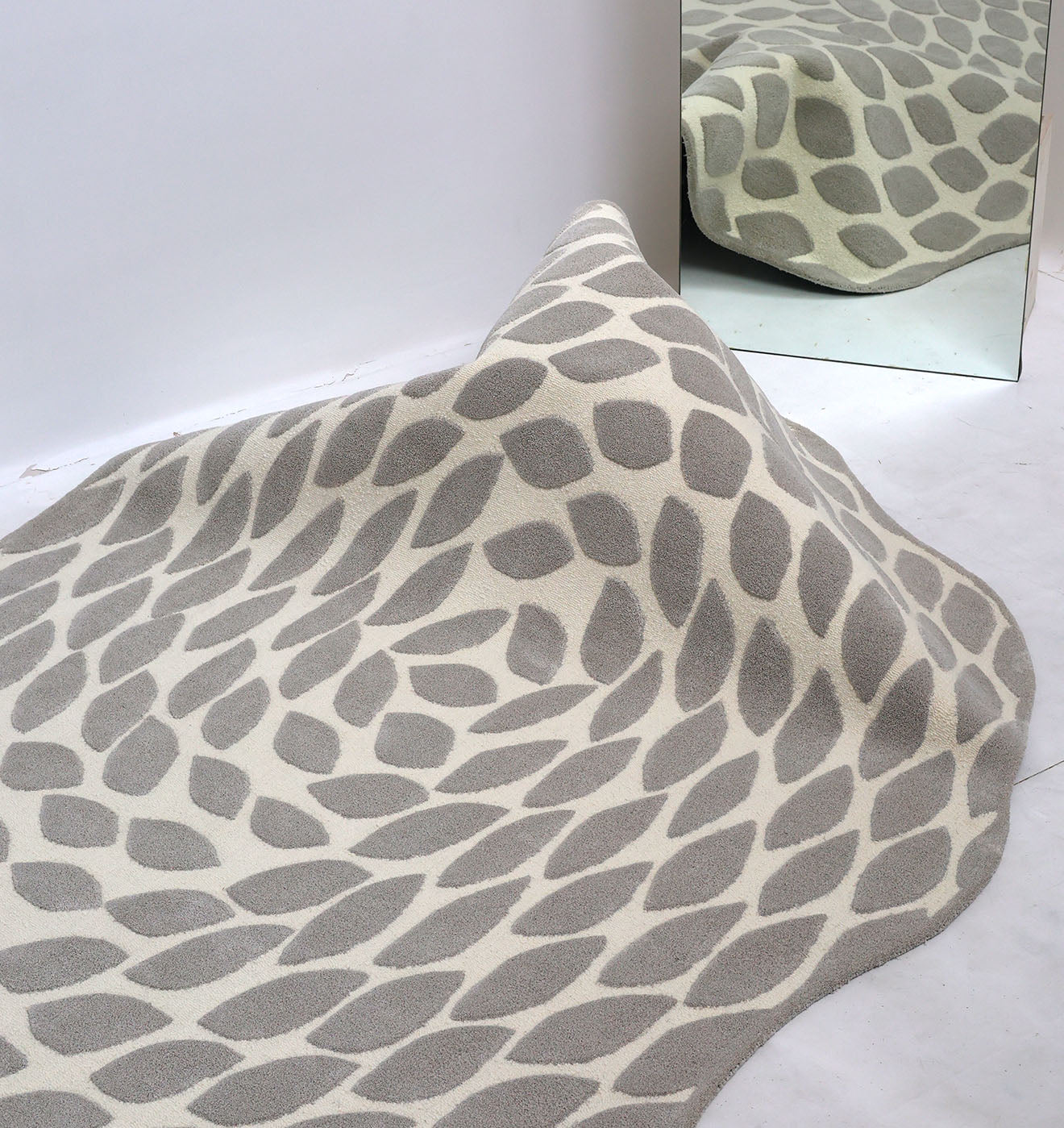The Handmade Shape Rug HTP-16 by RAMSHA HOME, featuring an irregular wool texture and gray leaf motif, is draped over a mound to form a hill shape, its reflection visible in a mirror placed at the corner.
