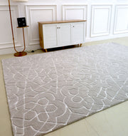 The Home Decor Area Rug HTP-17 by RAMSHA HOME, featuring a light color and subtle abstract floral design, lies on the floor of a minimalist room with white paneled walls, a compact wooden cabinet, and a gold floor lamp.