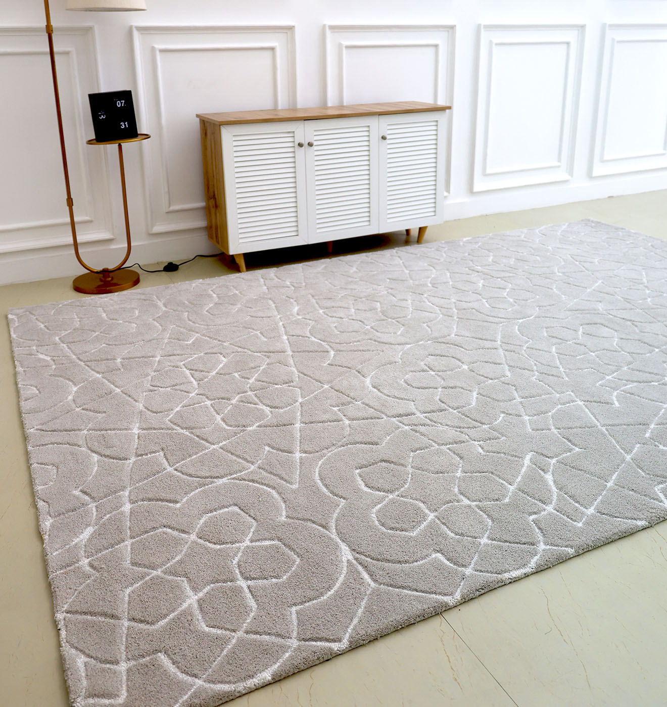 The Home Decor Area Rug HTP-17 by RAMSHA HOME, featuring a light color and subtle abstract floral design, lies on the floor of a minimalist room with white paneled walls, a compact wooden cabinet, and a gold floor lamp.
