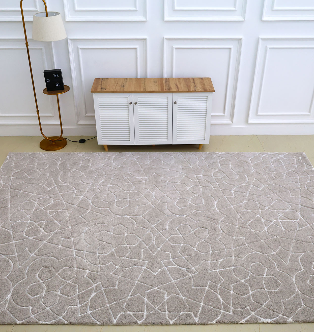 The RAMSHA HOME Home Decor Area Rug HTP-17, in light beige with a subtle geometric pattern, lies in front of a white cabinet with a wooden top, accompanied by a tall floor lamp and small black clock against a white paneled wall.