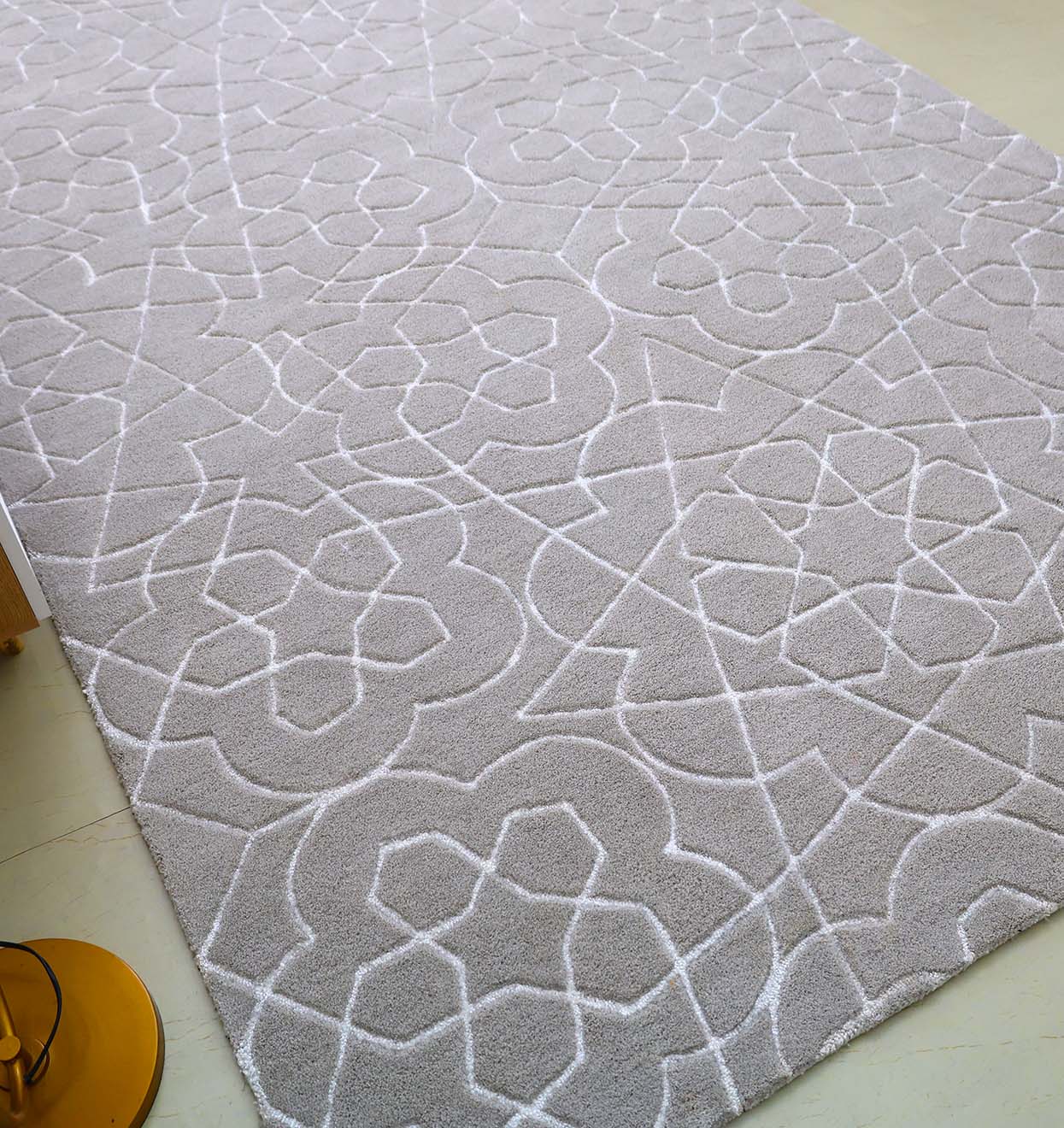 The RAMSHA HOME Home Decor Area Rug HTP-17 features a light grey hand-tufted design with a raised white geometric pattern of interlocking shapes and lines, placed on a light floor with a gold lamp base visible in the lower left corner.