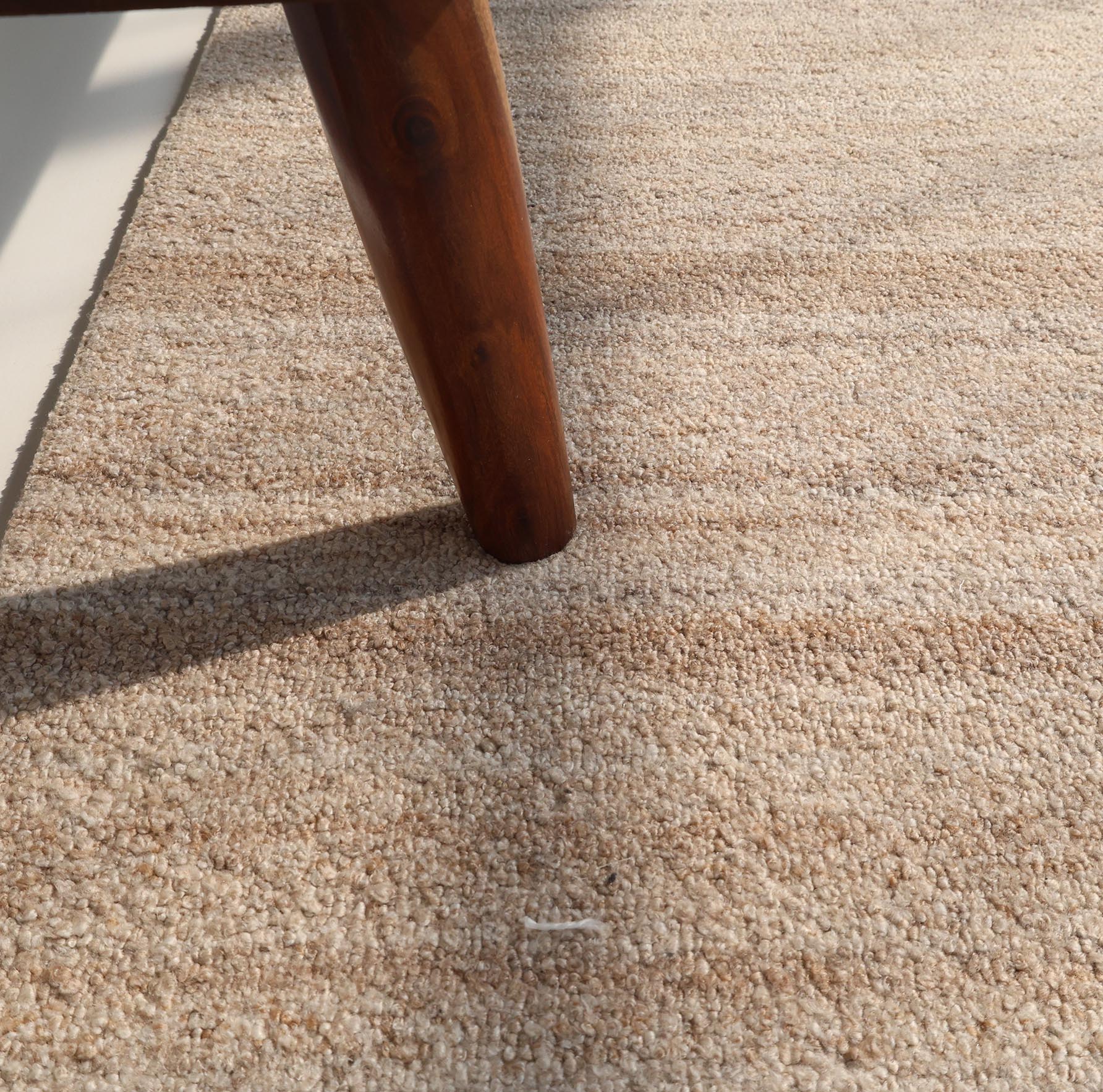 A wooden furniture leg rests on the Handmade Natural Wool Rug – Textured Beige Area Rug- HTP 27 by RAMSHA HOME. Sunlight casts shadows across the carpet, highlighting its texture, with a small white thread near the bottom center.