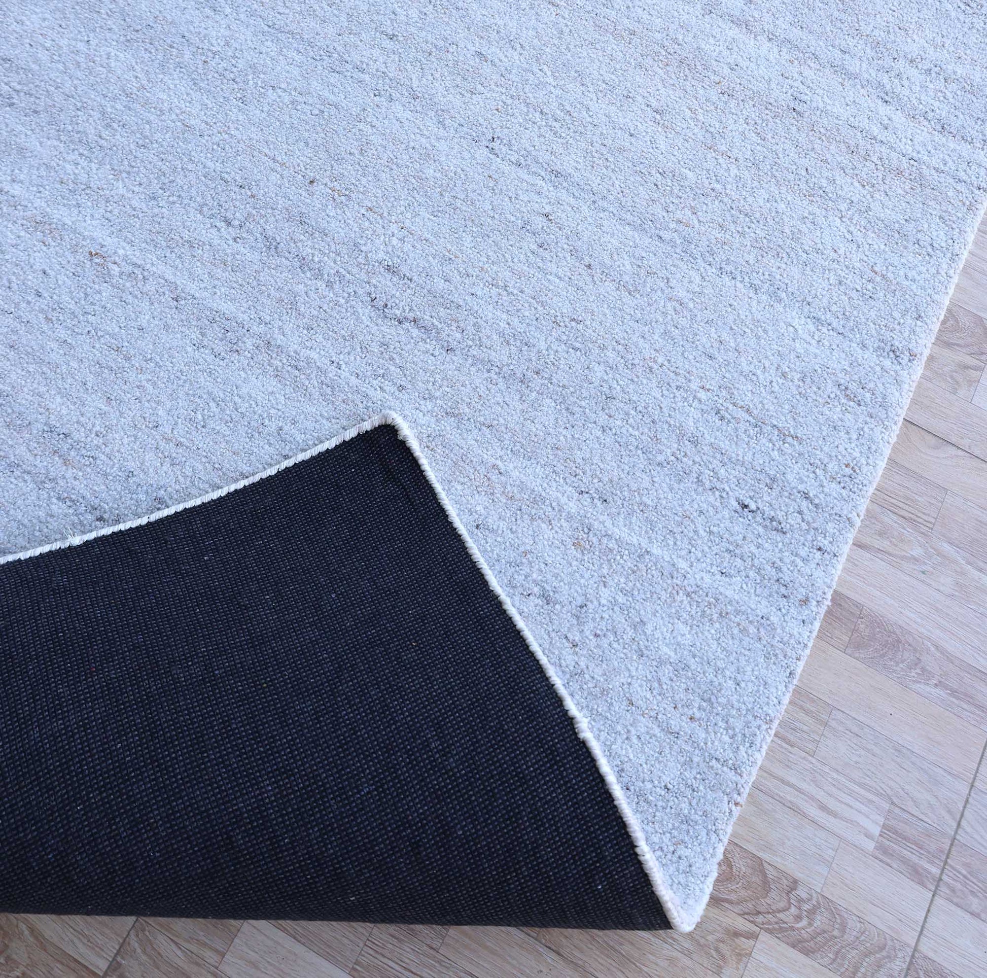 The Natural Ivory Handmade Wool Rug – Modern Luxury Area Rug HTP 28 by RAMSHA HOME, featuring a minimalist design, rests on a wooden floor with its corner lifted to show the black non-slip backing underneath.