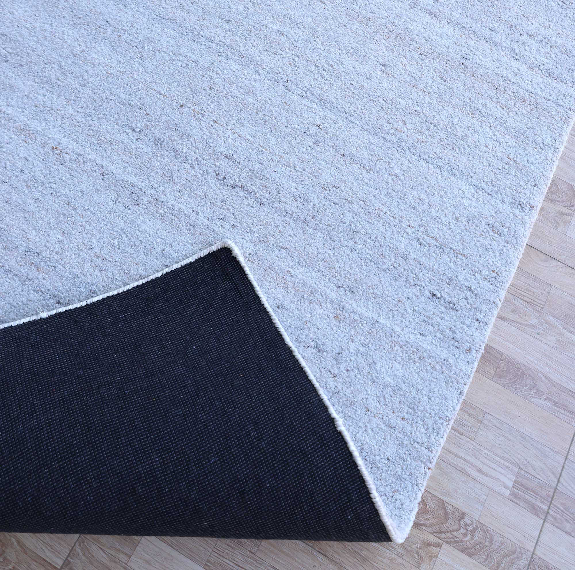 The Natural Ivory Handmade Wool Rug – Modern Luxury Area Rug HTP 28 by RAMSHA HOME, featuring a minimalist design, rests on a wooden floor with its corner lifted to show the black non-slip backing underneath.