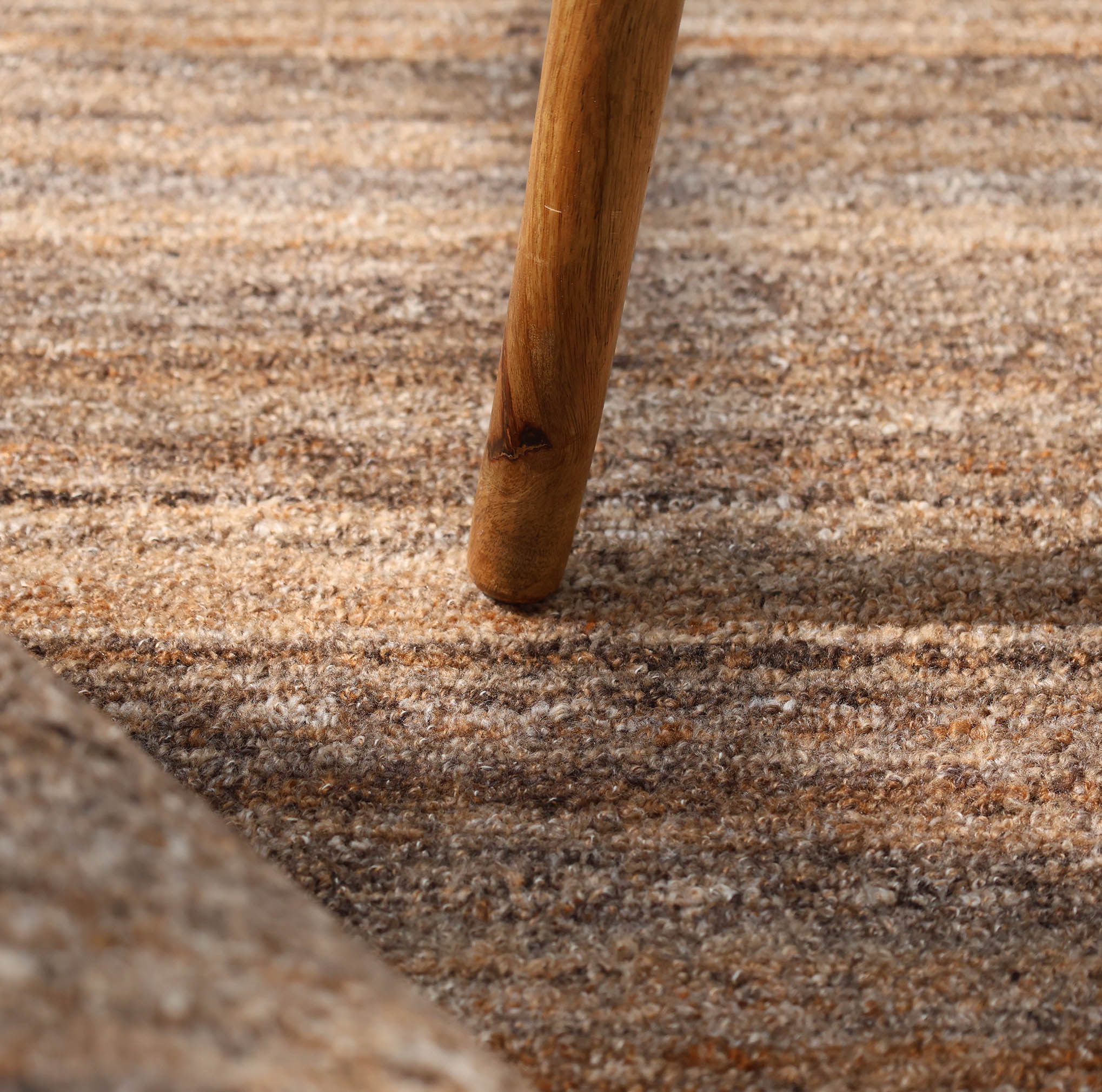 A close-up shows a wooden furniture leg on the RAMSHA HOME Handmade Earth Strata Wool Rug Brown Textured Area Rug HTP 29, highlighting the natural fibers and intricate textures of both materials at their contact point.