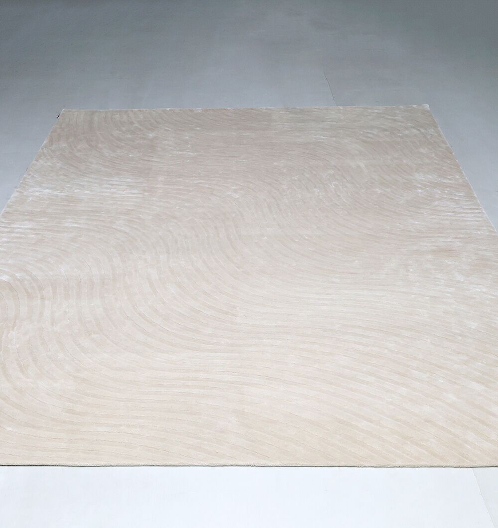 The Off White Handmade Rug - HTV 14 by RAMSHA HOME, a large square rug in light beige with subtle curved patterns, rests on a smooth light gray floor in a minimalistic setting.