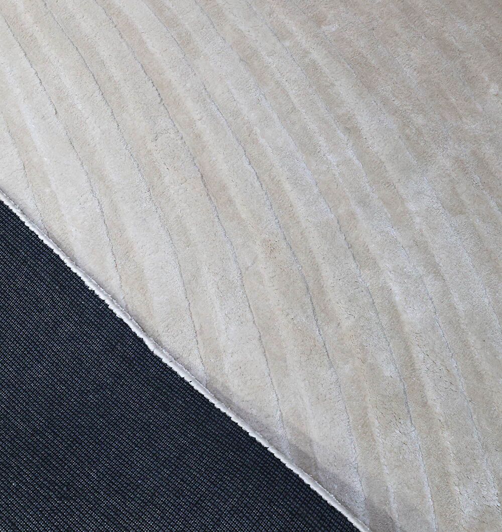 A close-up of the Off White Handmade Rug - HTV 14 by RAMSHA HOME, featuring a beige wavy pattern, partially lifted to show a black anti-slip rug pad underneath.
