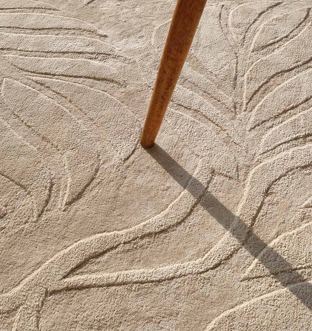A wooden table leg casts a shadow on the RAMSHA HOME Beige Color Indoor Handmade Area Rug - HTV 15, as sunlight highlights its textured, raised leafy pattern and intricate beige design.