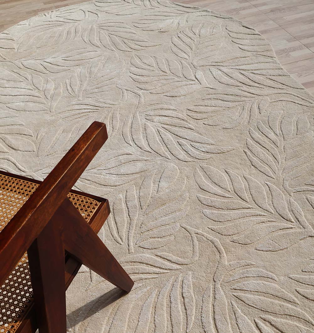 The RAMSHA HOME Beige Color Indoor Handmade Area Rug - HTV 15 with a raised leafy pattern lies on a light wood floor. The lower left corner of the image shows part of a wooden chair with a woven cane backrest.