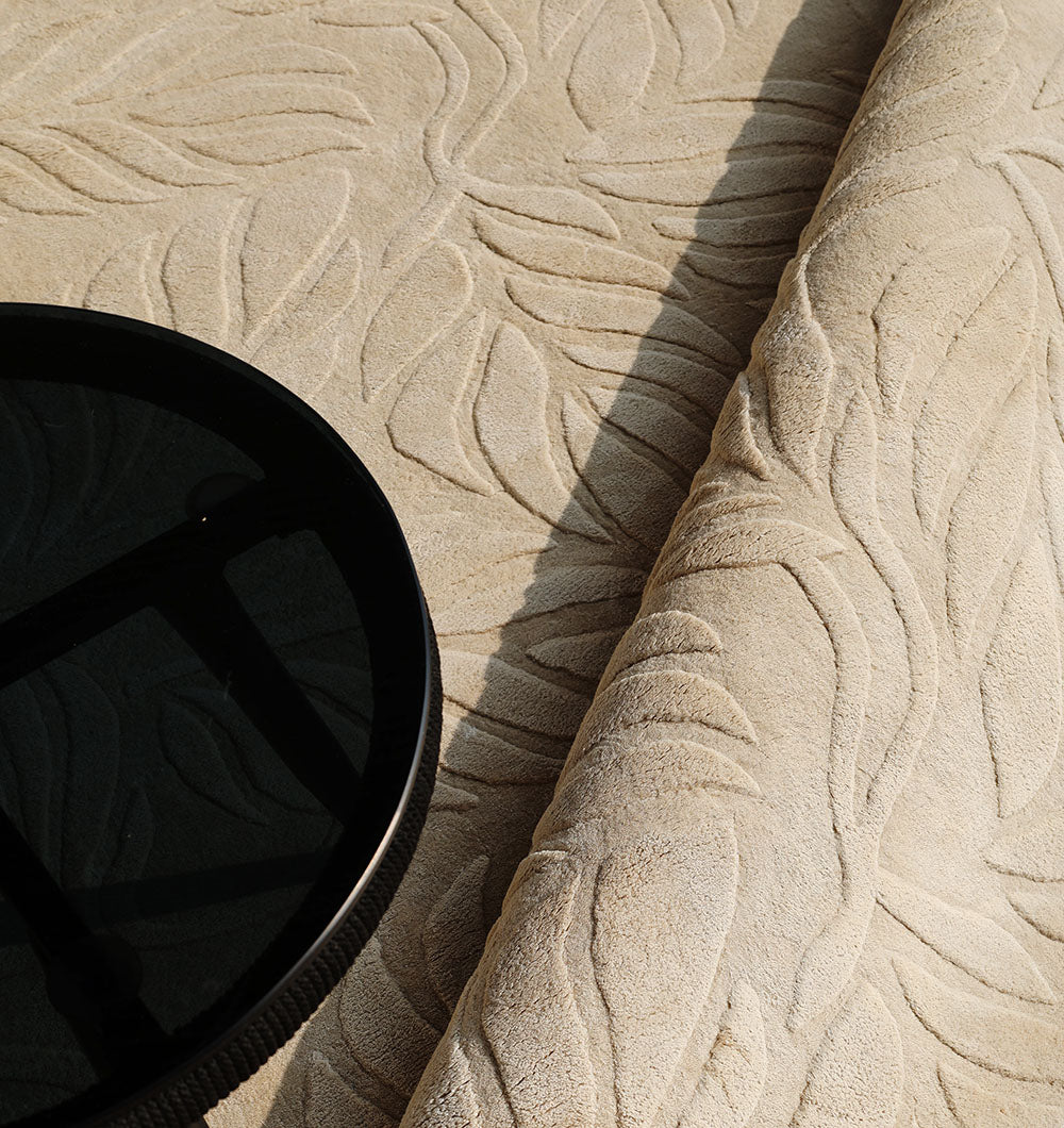 The RAMSHA HOME Beige Color Indoor Handmade Area Rug - HTV 15, featuring a raised leaf-like pattern, is partially rolled next to a round black glass-topped table and casts a soft shadow on the textured floor.