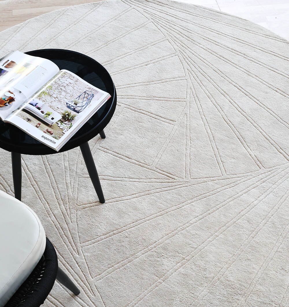A round black side table with an open magazine rests on the RAMSHA HOME Home Decor Handmade Rug - HTV 16 (200cm), a light beige handtufted wool rug with geometric patterns; a white chair is also partly visible.