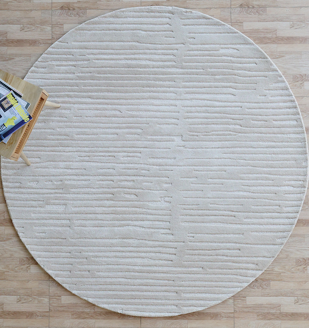 The RAMSHA HOME Decor Handmade Area Rug - HTV 17, round and cream-colored with subtle horizontal lines, is displayed on a light wood floor. A small side table with books and a magazine appears partially in the top left corner.